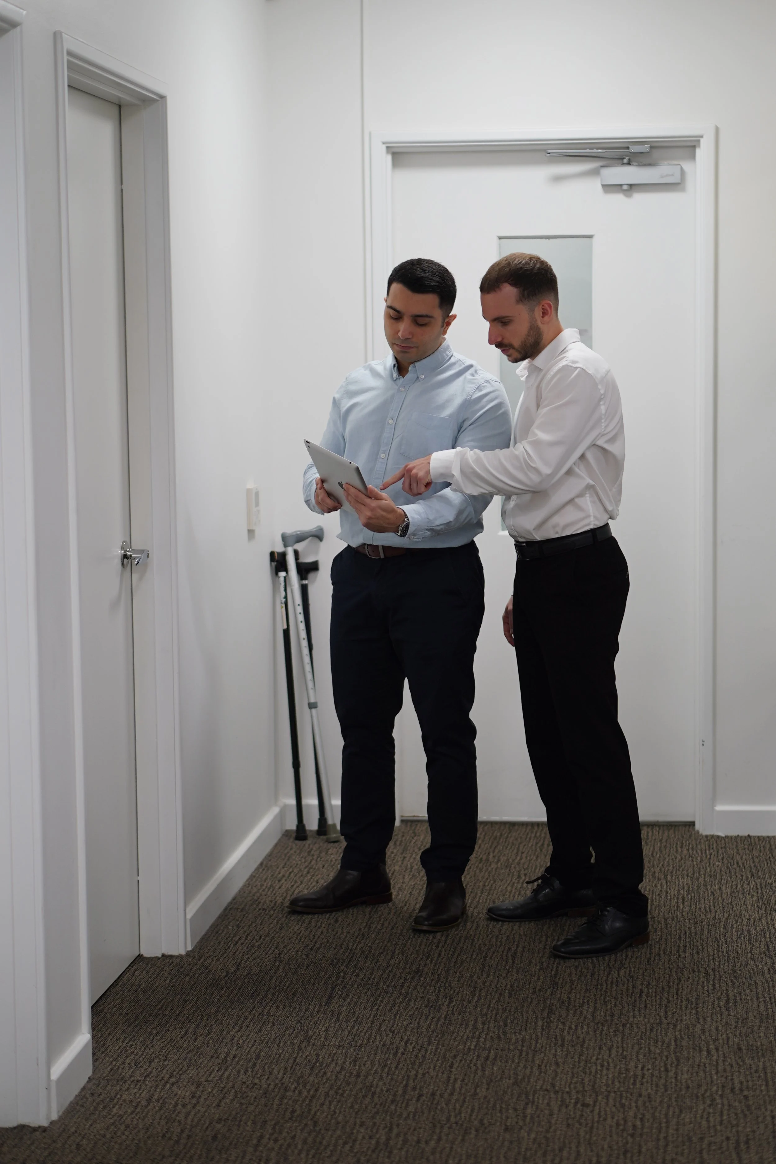 Two men in dress shirts and black pants standing in a hallway, looking at a tablet. One man is pointing at the tablet, and a cane is leaning against the wall nearby.