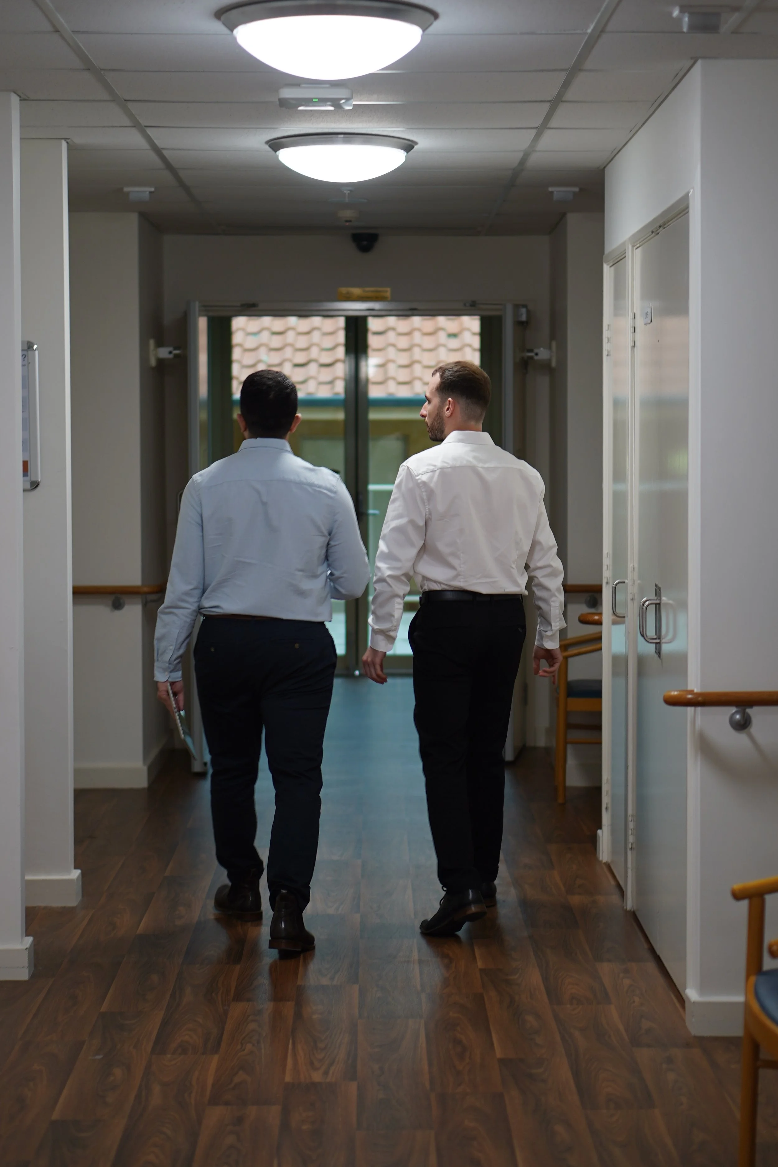 Two men in business attire walking down a hospital corridor, engaged in conversation. One is holding a clipboard and the other has a beard and is looking to the side. The corridor has wooden flooring, ceiling lights, and handrails on the walls.