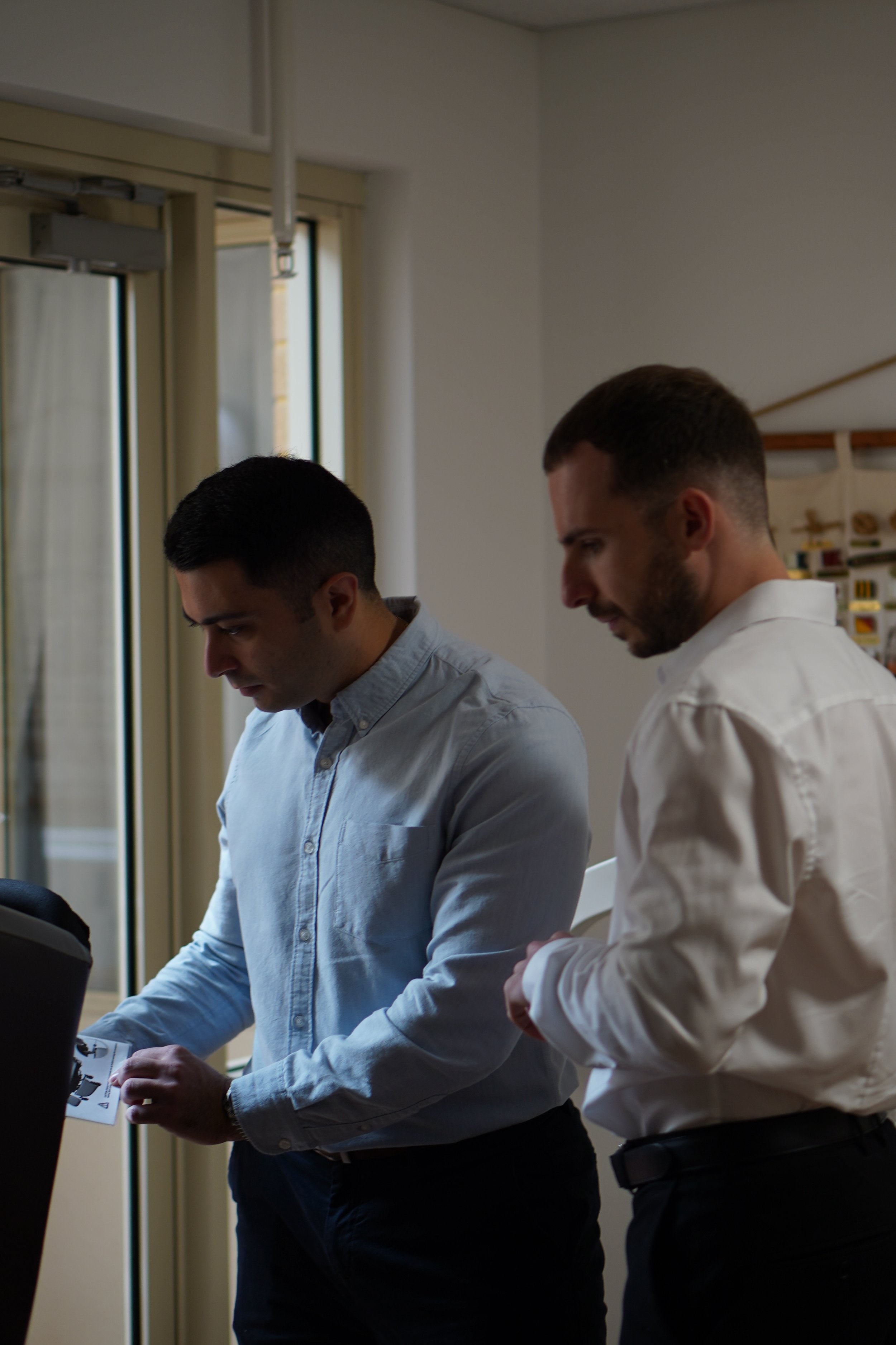 Two men standing indoors, one looking down at an object and the other looking over his shoulder, with a window and a bookshelf in the background.