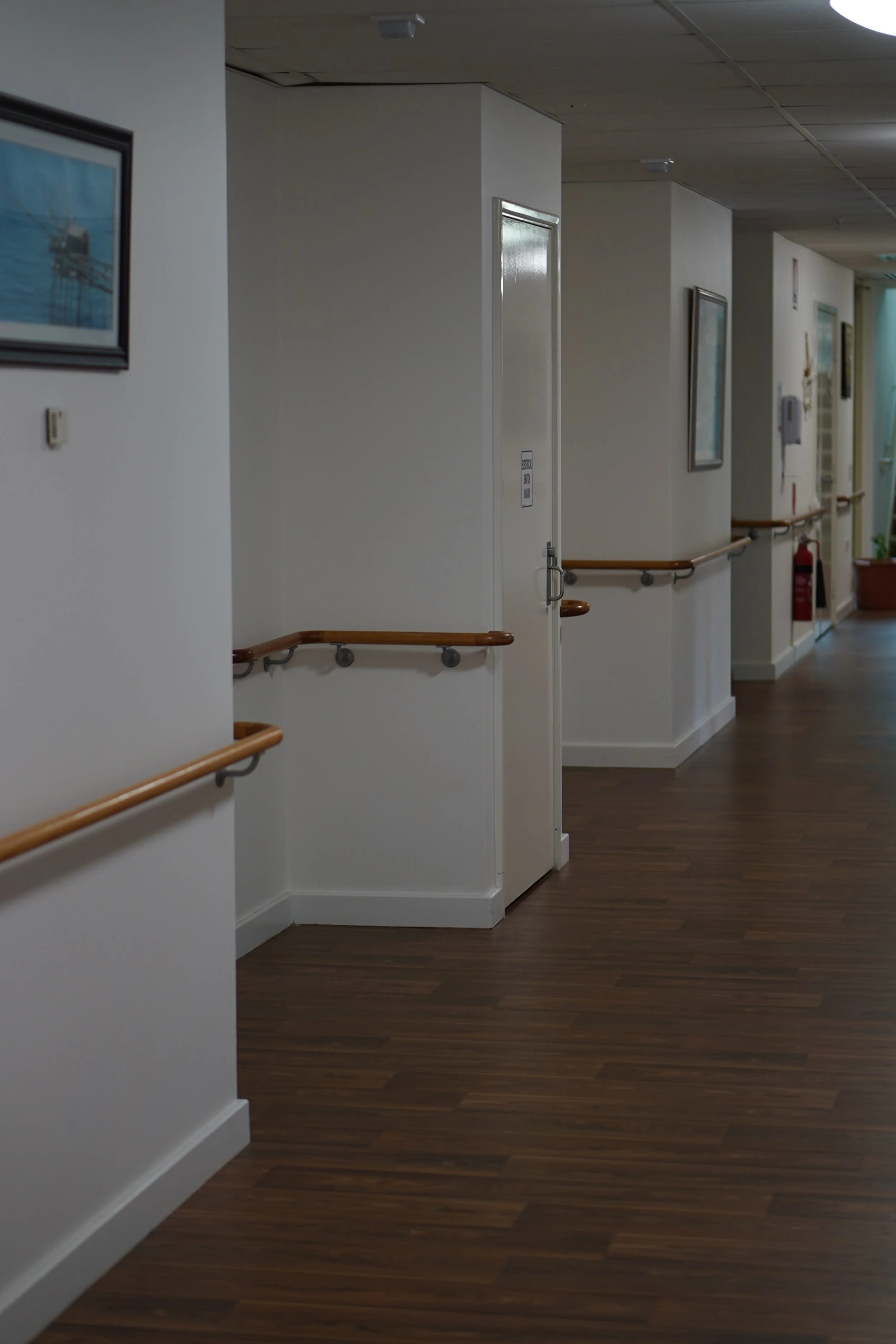 A hallway in a healthcare or care facility with white walls, wooden handrails, framed pictures, a fire extinguisher, and a door with a sign on it.