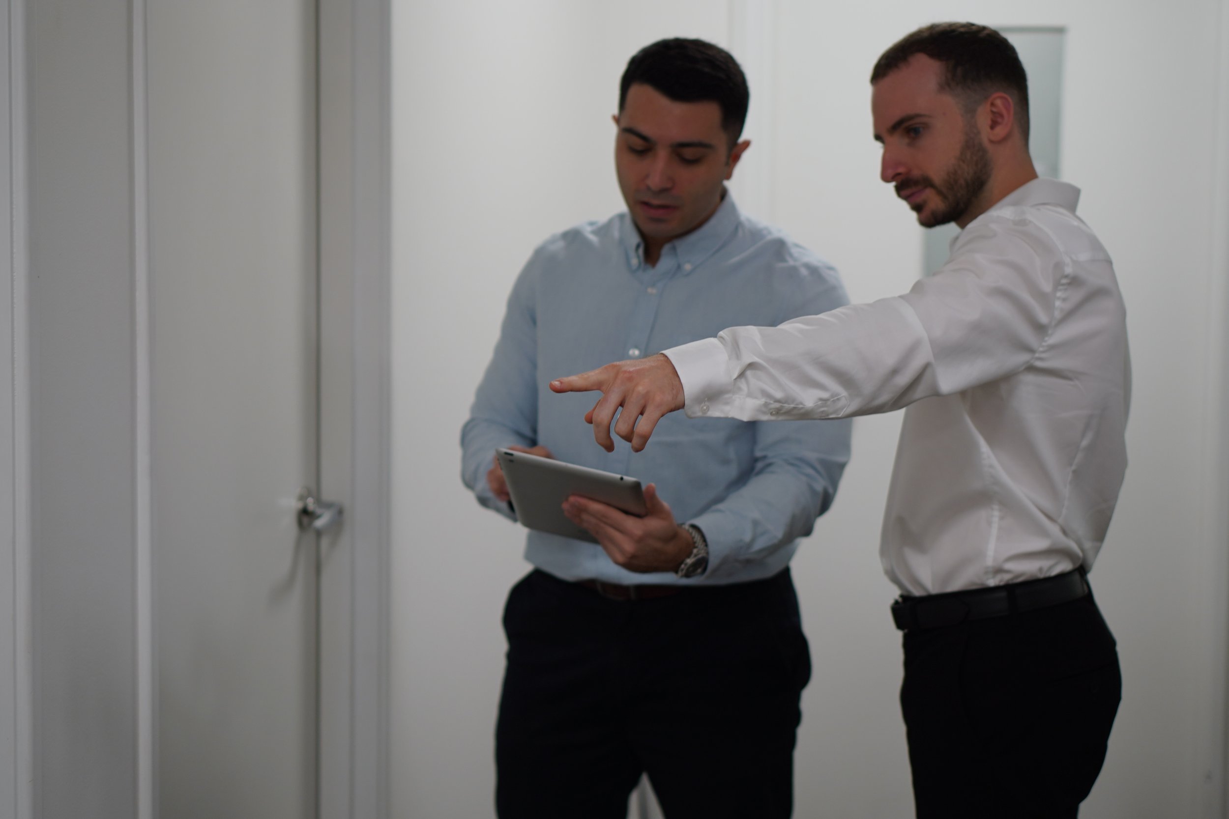 Two men are standing in a corridor, one is holding a tablet and the other is pointing at it, both dressed in business casual attire.