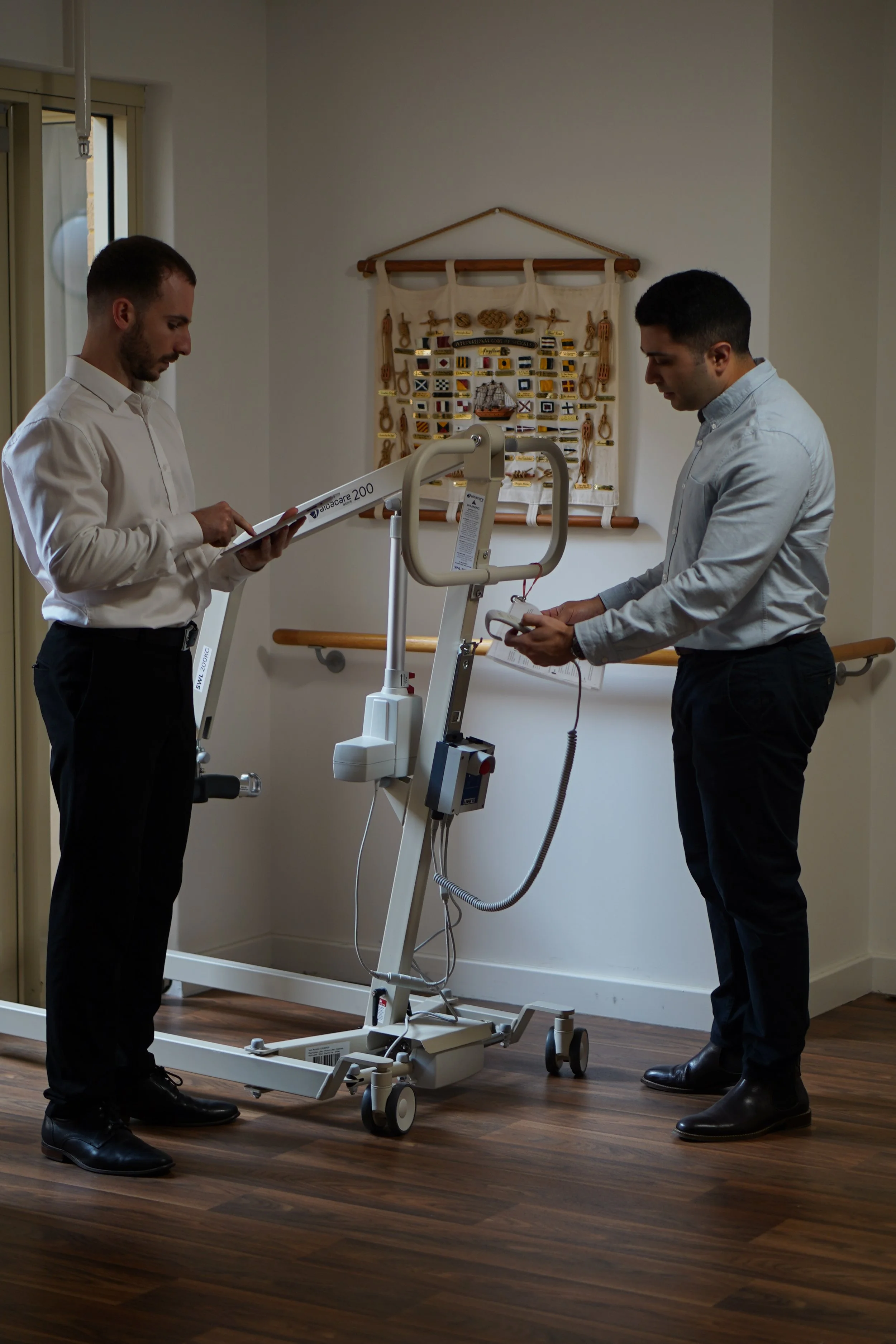 Two men in business attire are standing near a medical rehabilitation device in a room. One man is reading a clipboard, and the other is holding a device connected to the machine. The room has framed nautical-themed wall decor and wooden flooring.