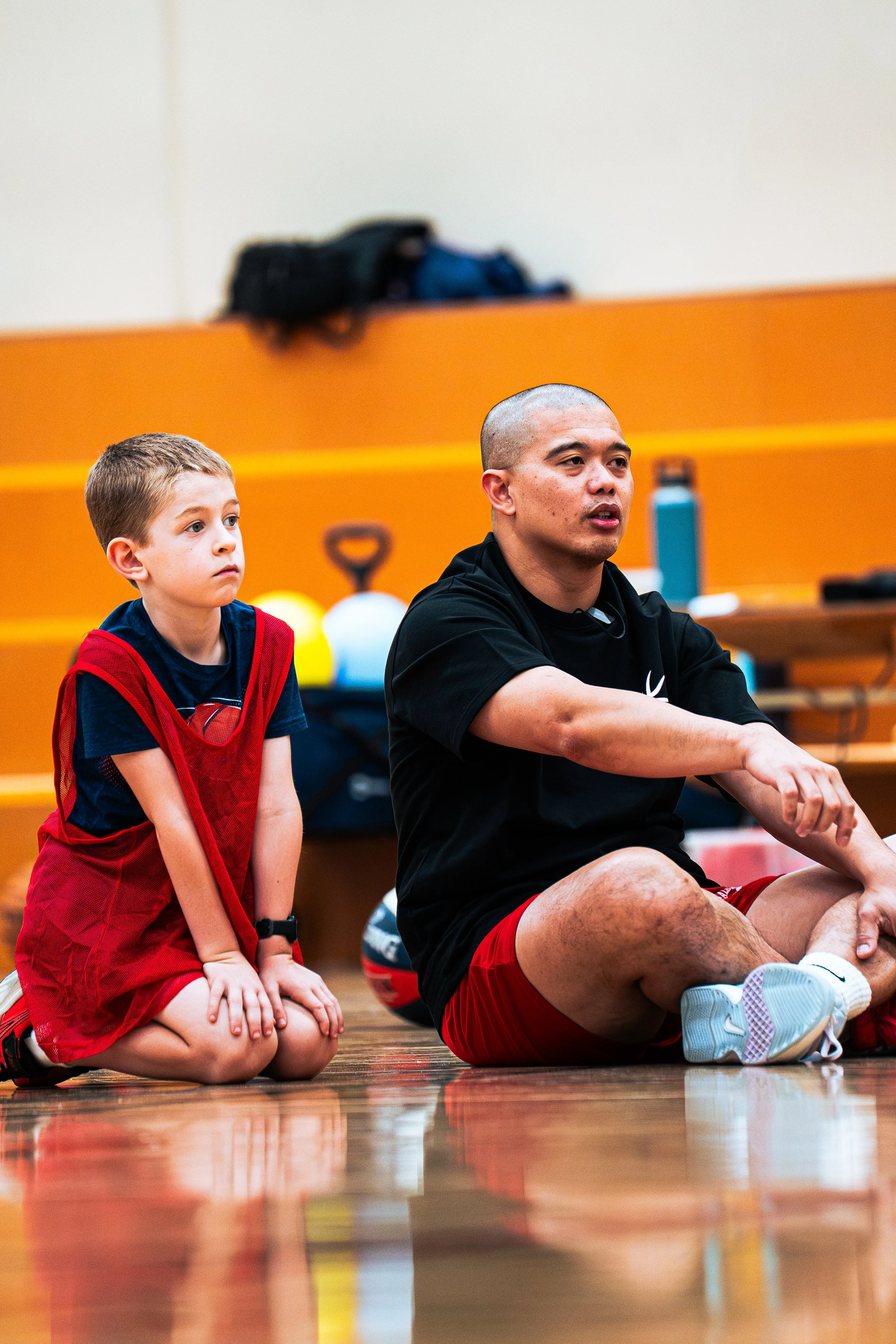 A young boy and an adult man sitting on a gymnasium floor, both in a kneeling position, with the boy wearing a red sports jersey and the man sitting cross-legged in athletic clothing. They appear to be participating in or watching a sporting activity. In the background, there are gym equipment and backpacks on a yellow bench against a beige wall.