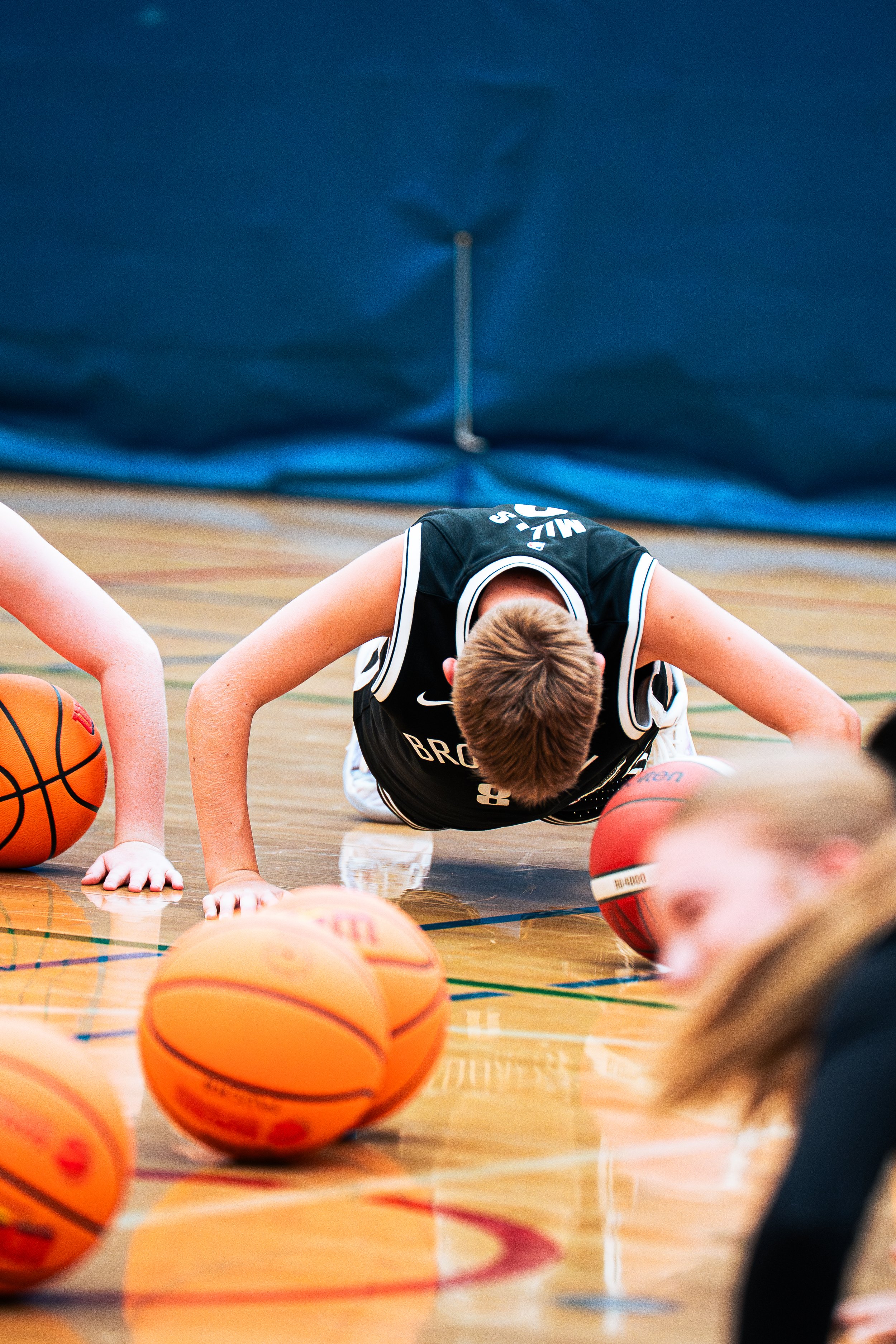 A young basketball player in a black jersey doing a push-up on a basketball court, surrounded by multiple basketballs.