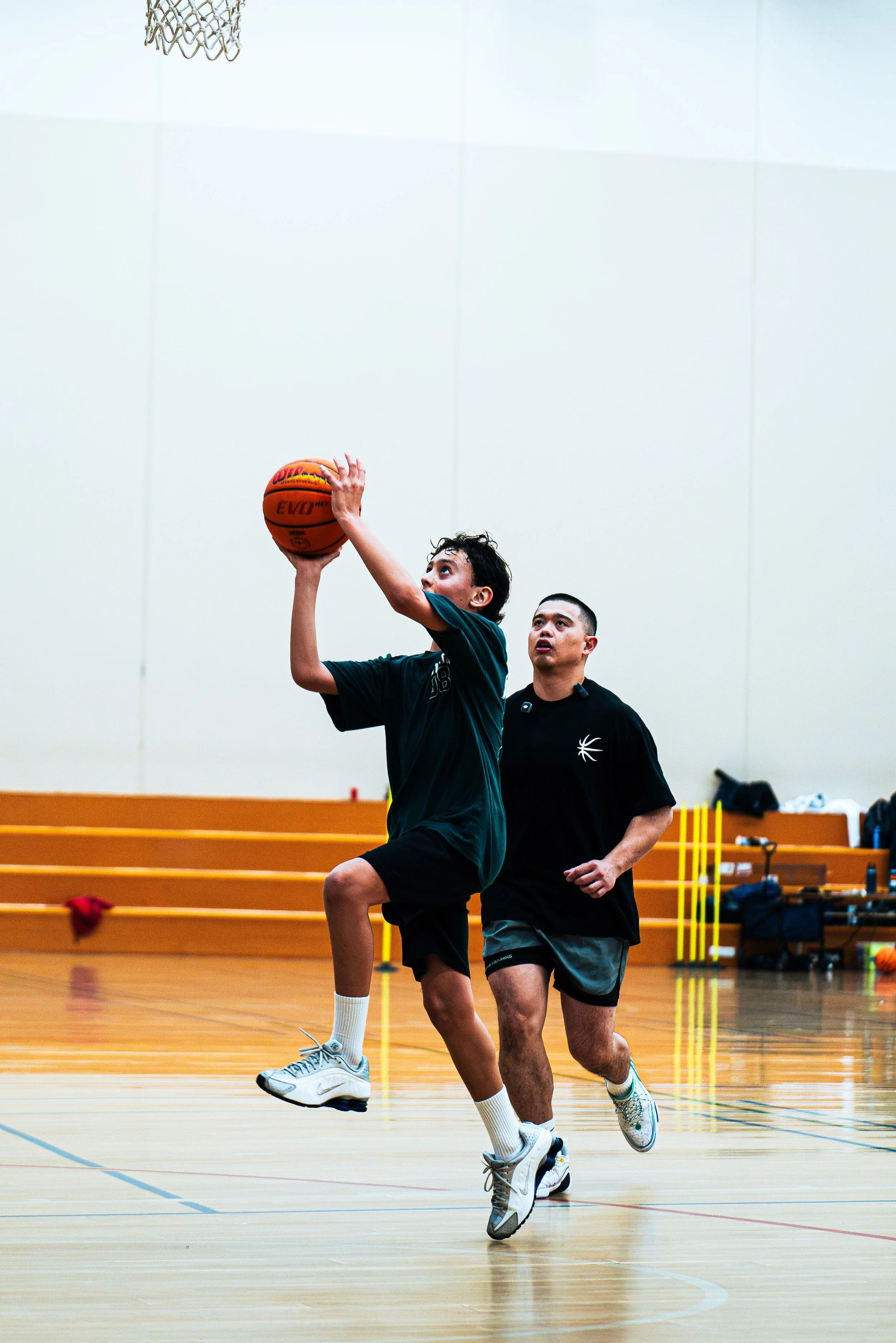 Two young men playing basketball in an indoor gym, one jumping to shoot the ball towards the hoop, while the other closely follows, with a wooden floor and gym equipment in the background.
