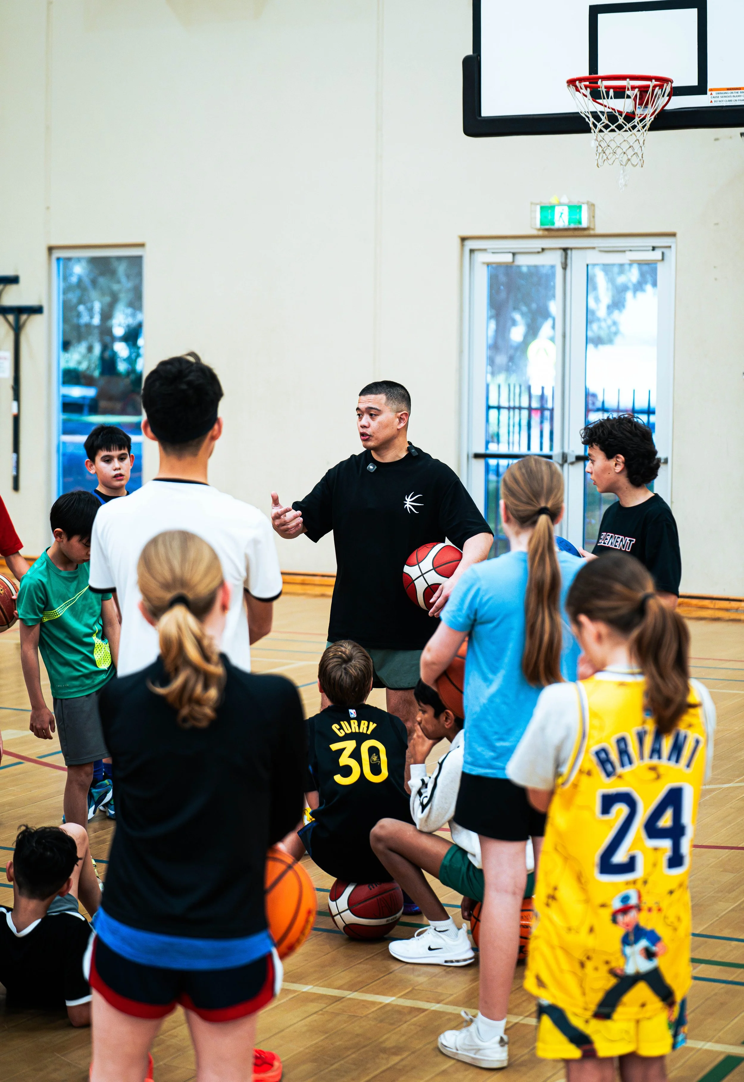 A group of children gathered around a coach in a gymnasium with basketballs, listening to instructions during a basketball clinic.