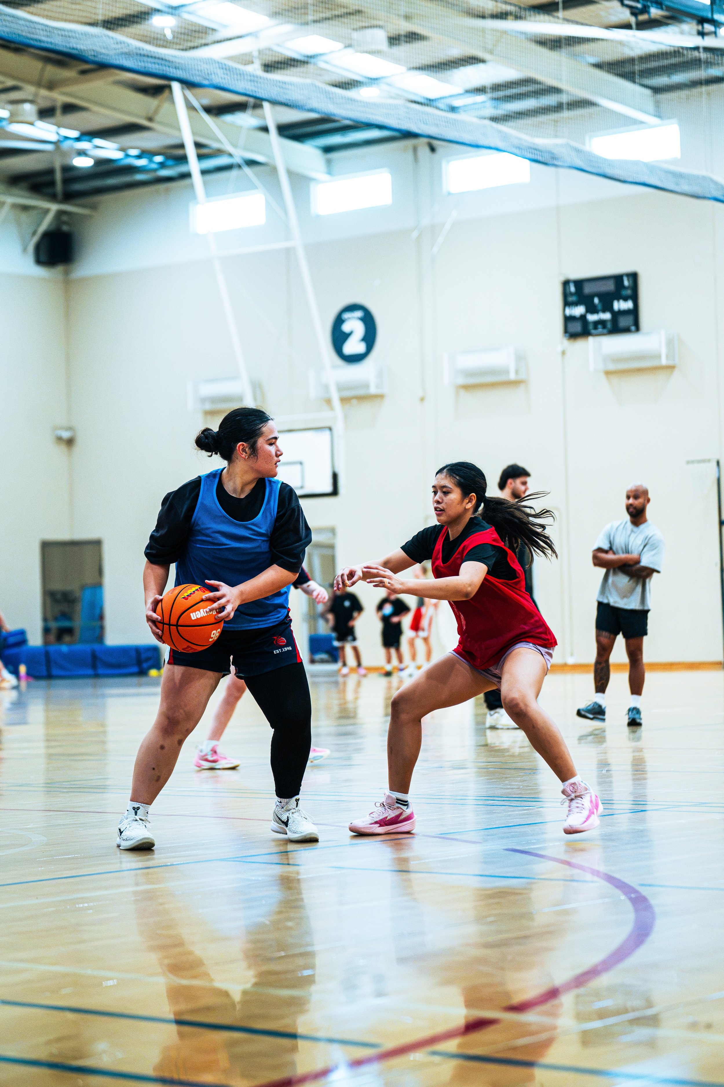 Two female basketball players are playing defense and offense on an indoor basketball court while a man is observing in the background.