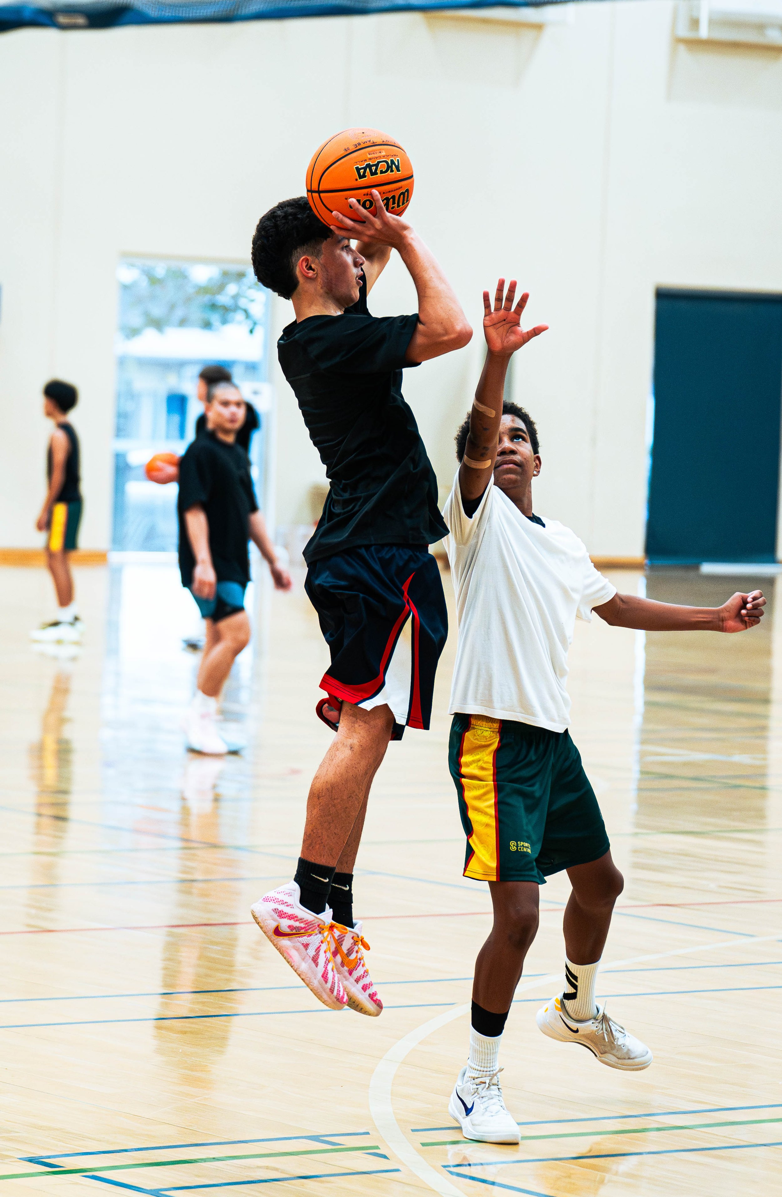 Two young boys playing basketball indoors, one jumping to make a shot while the other tries to block. Other children are visible in the background on a gymnasium court.