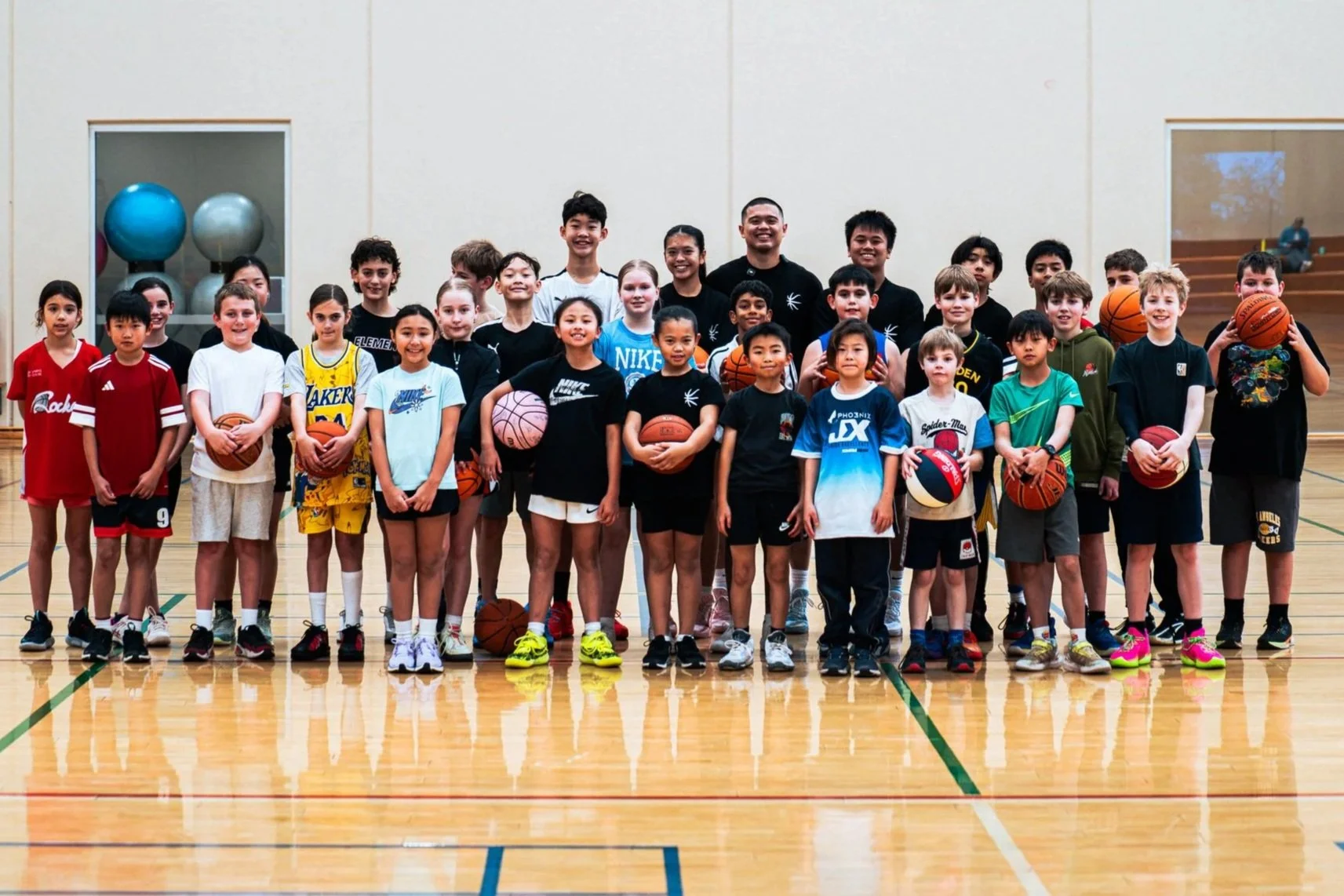 A group of children and a few adults standing in a gymnasium holding basketballs, posing for a group photo. The children are dressed in sports attire, and the background shows a gym with wooden flooring and some sports equipment.