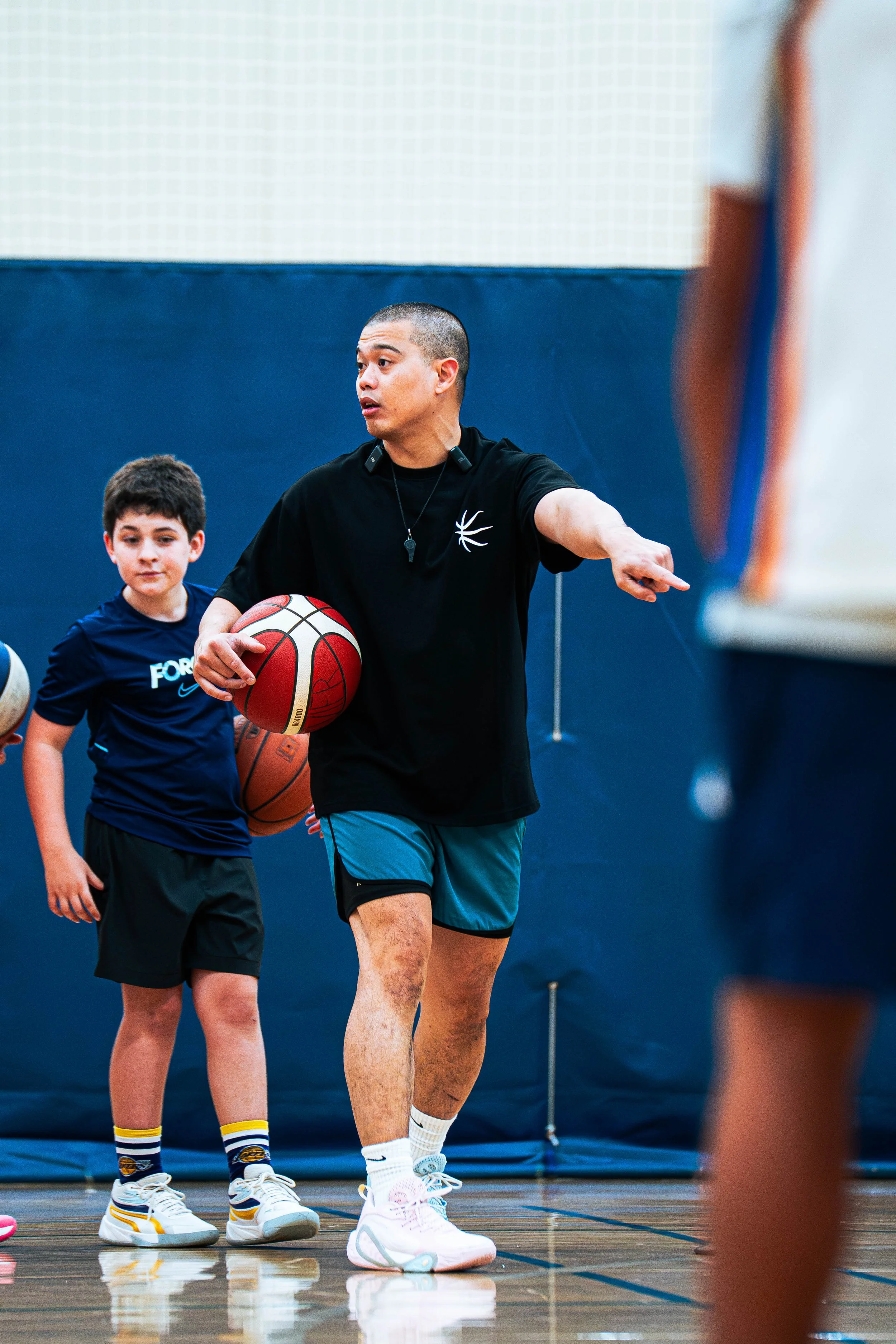 A basketball coach instructs young players during a basketball practice session in an indoor gym. The coach is holding a basketball and is pointing, while a boy in a blue jersey stands nearby holding another basketball, listening attentively.