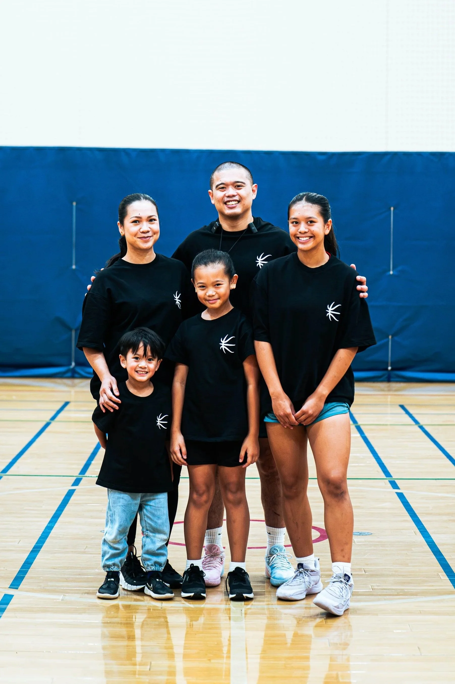 Group of five people, including three young women, a girl, and a boy, in a gymnasium, wearing matching black t-shirts with a white logo, smiling and posing for the photo.