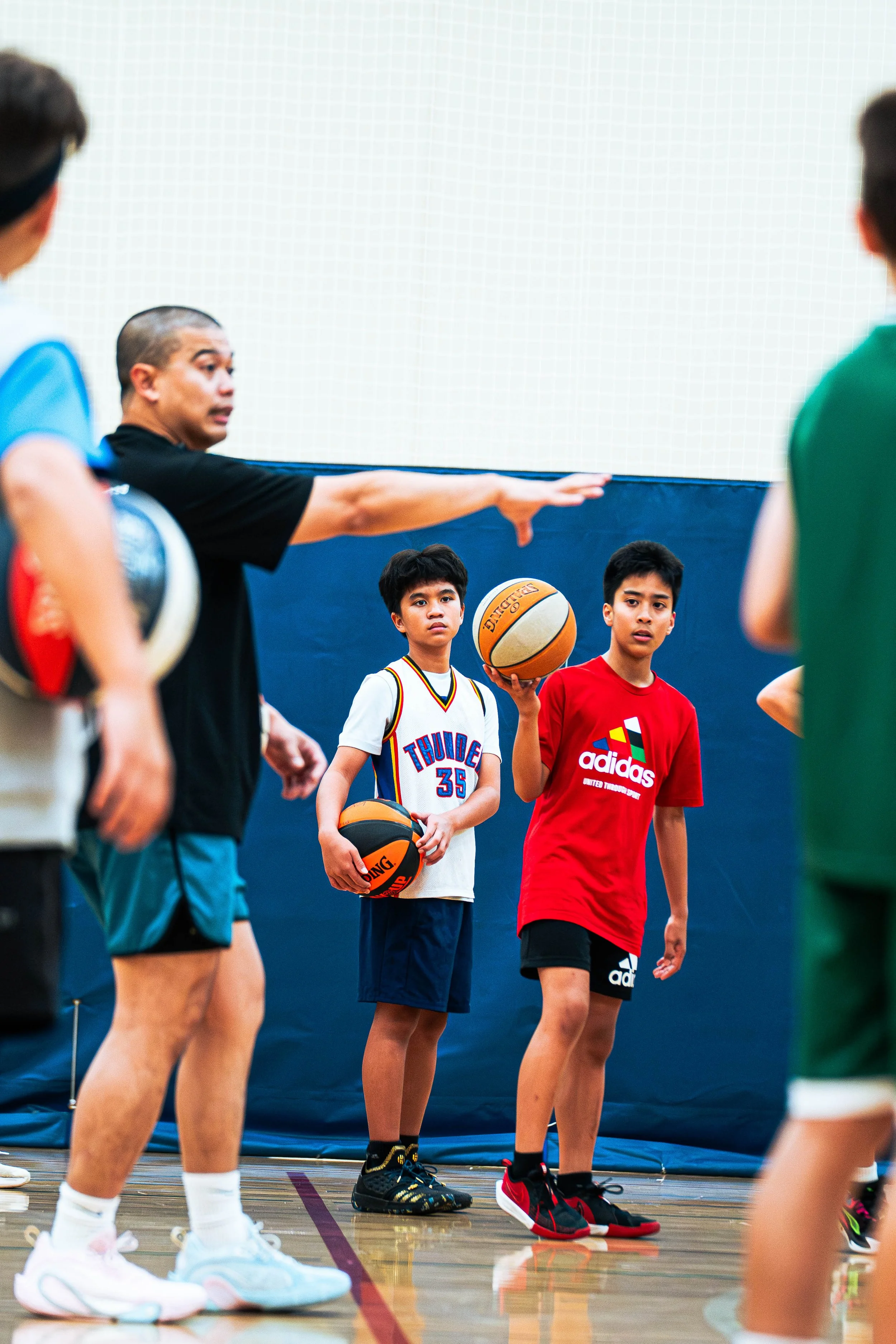 Boys in a basketball gym during a drill, with a coach demonstrating a move, wearing casual sportswear, and holding a basketball.