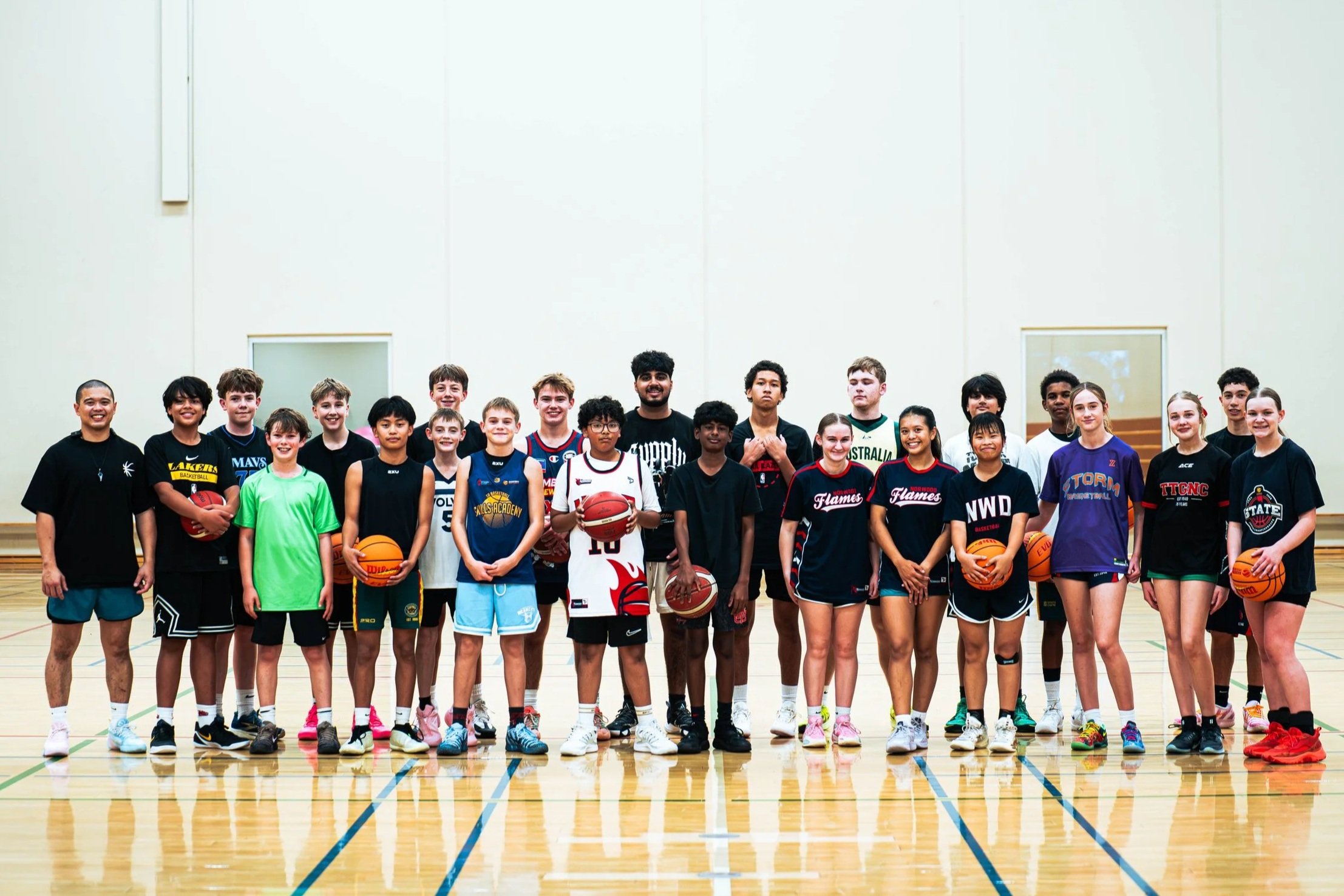 Group of children and teenagers in basketball uniforms posing for a team photo in an indoor gymnasium, some holding basketballs.