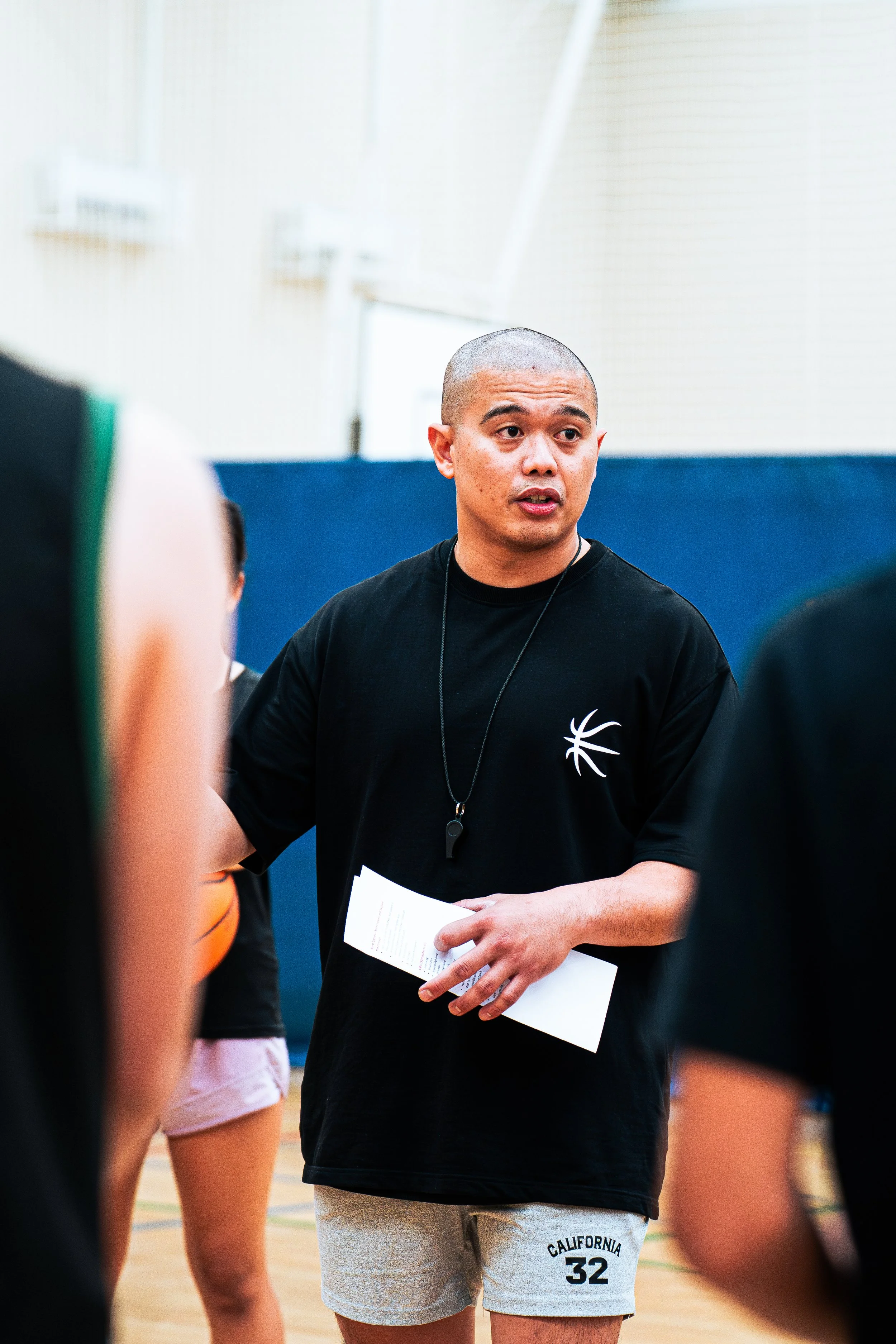 A man with a shaved head and light brown skin is standing in a gymnasium, holding a piece of paper, and talking to a group of people whose bodies are partially visible. He is wearing a black t-shirt with a white basketball logo on it and light gray shorts with the phrase 'California 32' on the leg.