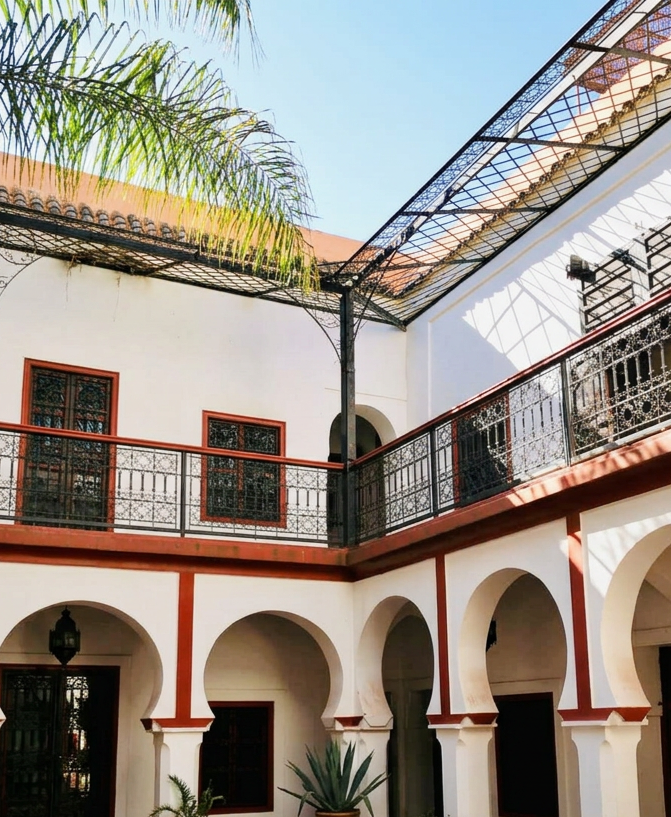 Interior courtyard of a building with white walls, arched openings, decorative iron balconies, a red metal roof, and a metal and glass roof structure providing partial shade. There are potted plants and greenery visible.