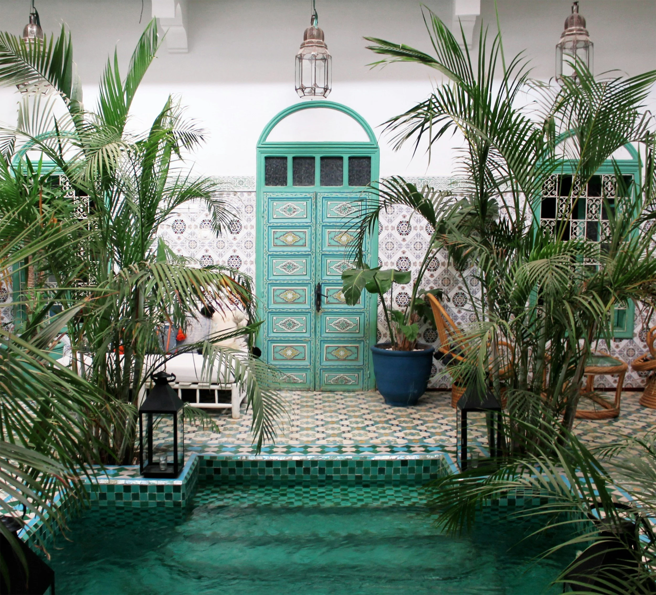 A decorative indoor courtyard with lush green plants surrounding a small pool with green tiles, a turquoise patterned door, and hanging lanterns, with tiled floors and intricate wall designs.
