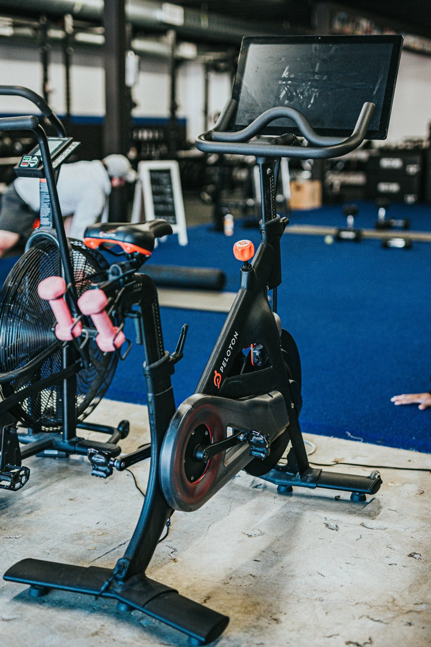 Indoor gym with stationary exercise bike, pink dumbbells, and a fan on a metal stand in the foreground. A person in workout attire and a white cap is bending over in the background, near a blackboard and other gym equipment.