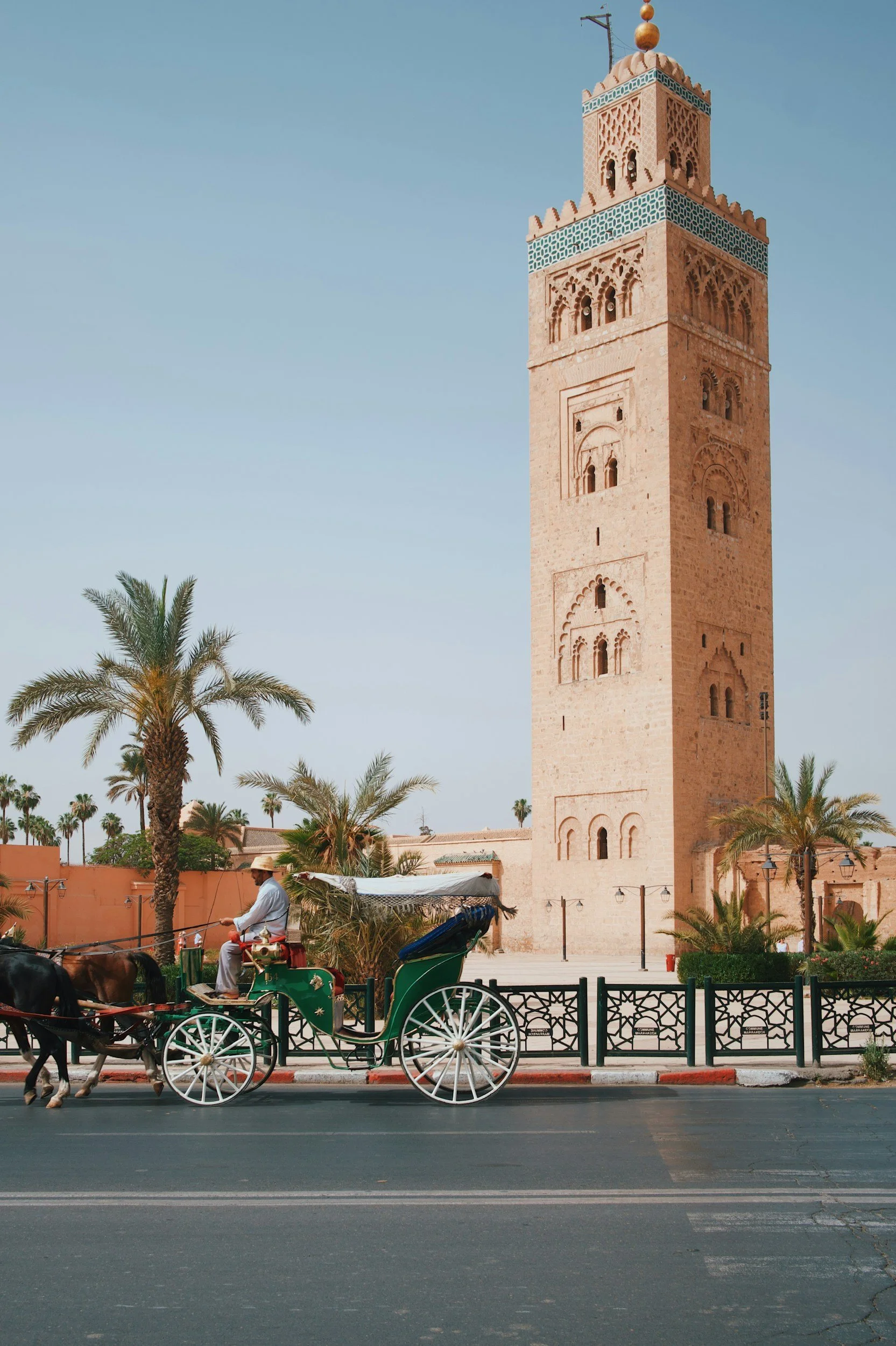 A street scene in a North African city featuring a tall, ornate minaret of a mosque, palm trees, and a horse-drawn carriage with a man dressed in white. The sky is clear and blue.