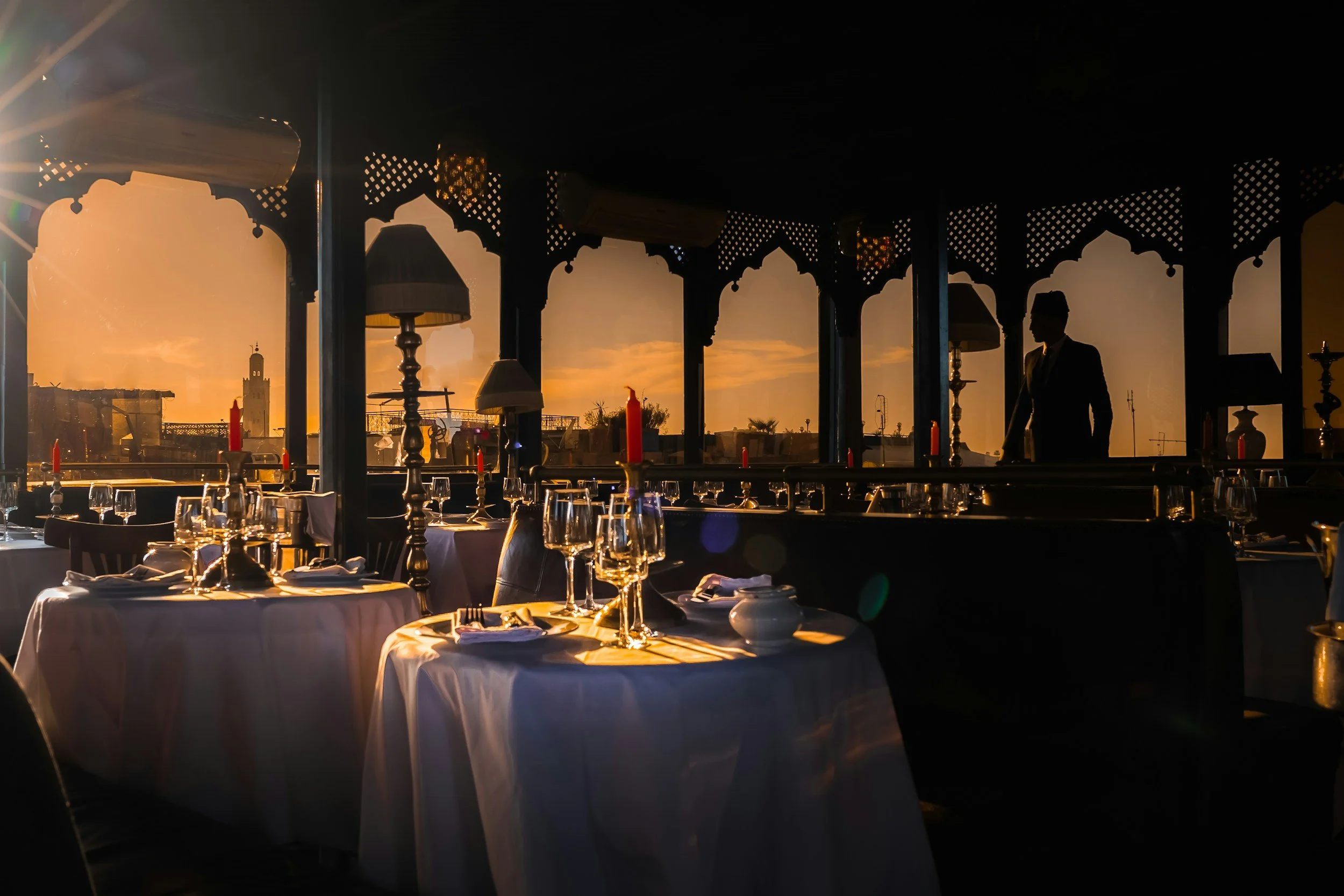 Interior of an elegant restaurant with white tablecloths, candles, wine glasses, and silverware, featuring a view of the sunset outside with a silhouette of a man in a suit, decorative lamps, and cityscape in the background.