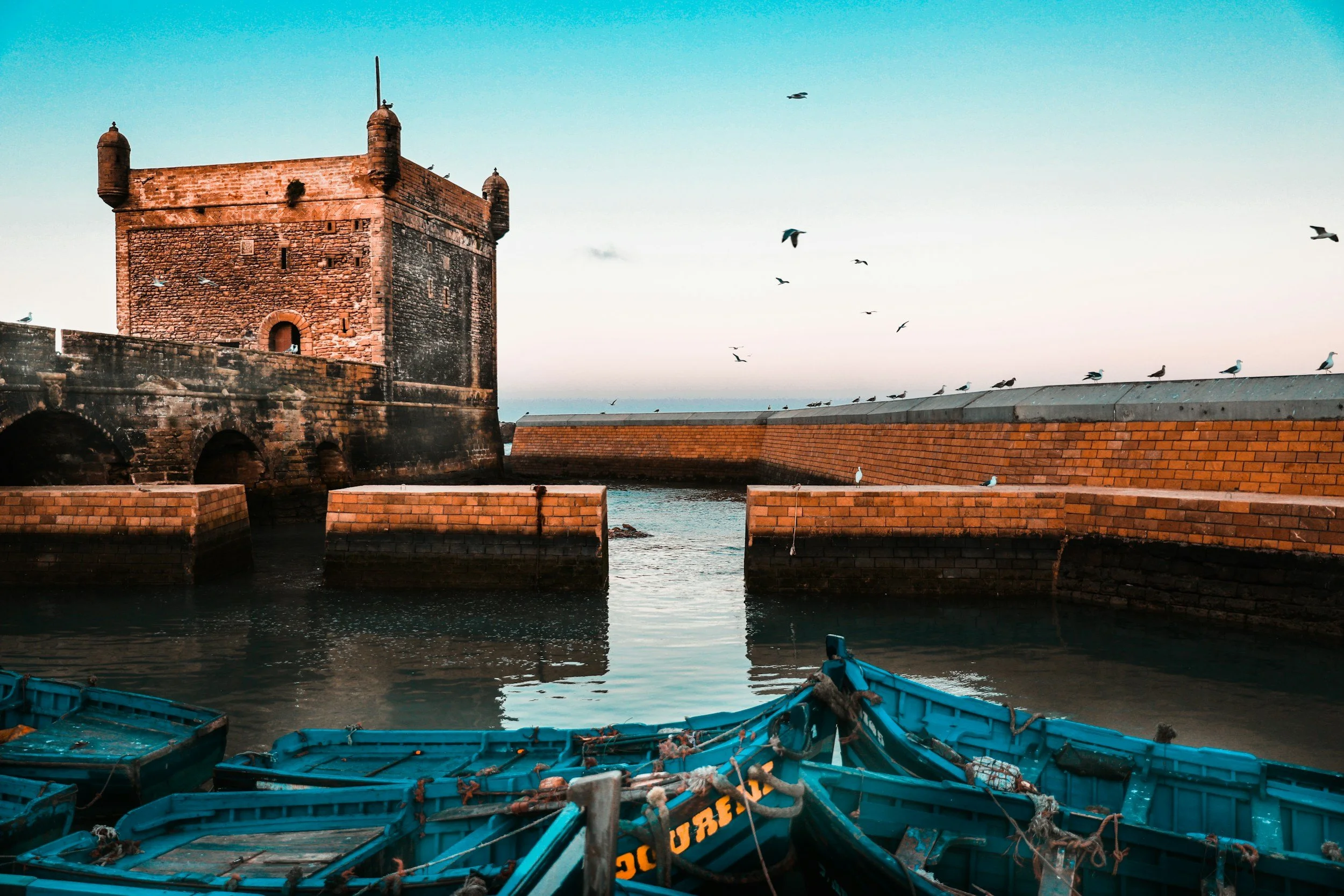 Historic stone fortress by the water with seagulls flying overhead and boats in the foreground.