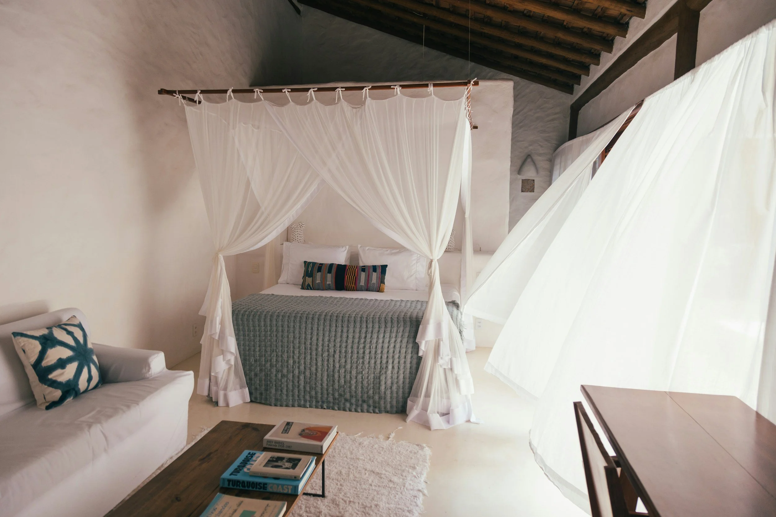 A cozy bedroom featuring a white canopy bed with sheer curtains, a white sofa with patterned pillows, a wooden coffee table with books, and a rustic ceiling with exposed wooden beams.