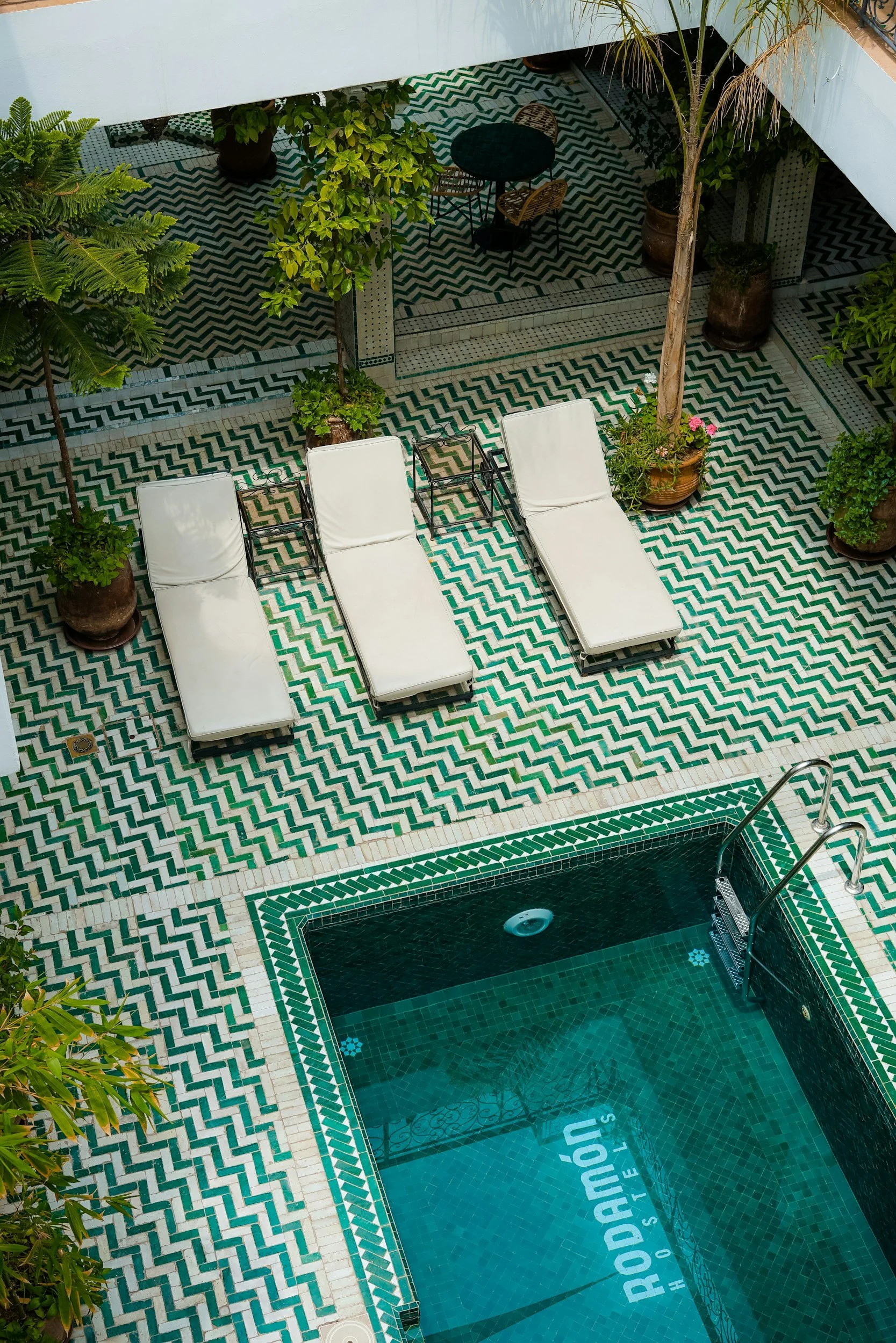 A view of a swimming pool with clear water and a pool ladder at the bottom right corner. The pool is surrounded by a decorative tile border with green and white patterns. Adjacent to the pool, there are three white lounge chairs with a small glass ta
