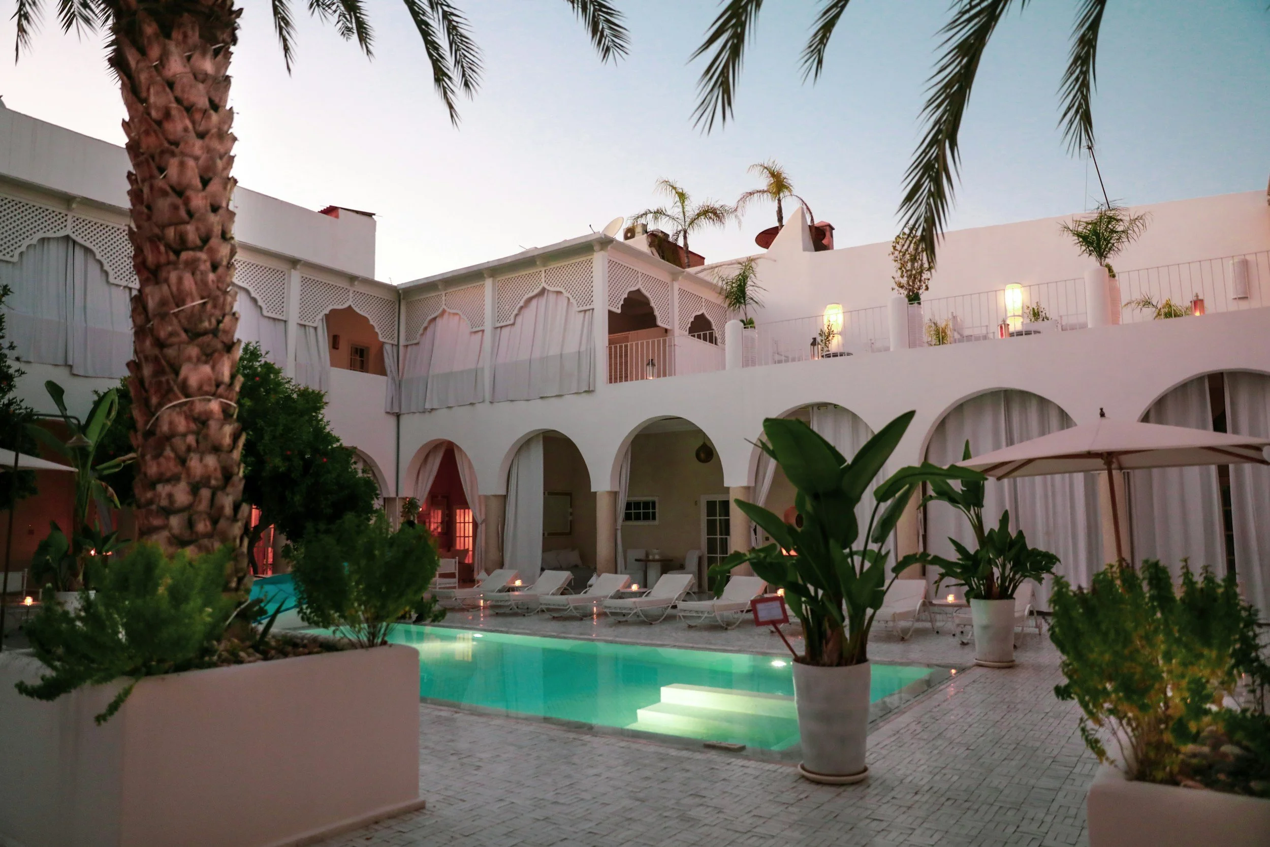 A nighttime view of a white hotel courtyard with a swimming pool, lounge chairs, and potted plants, including palm trees. The hotel has arched windows and balconies with sheer curtains, and there are lights illuminating the area.