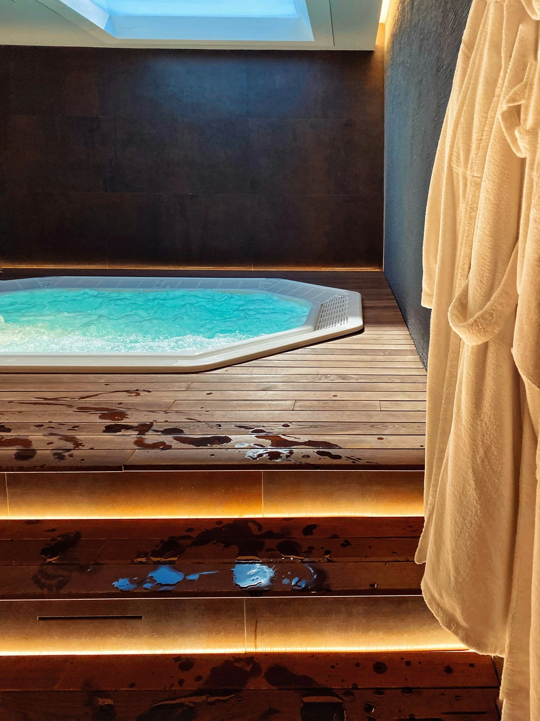A wooden floor with water puddles, a white hot tub filled with blue water, dark walls, and a beige curtain on the right side.
