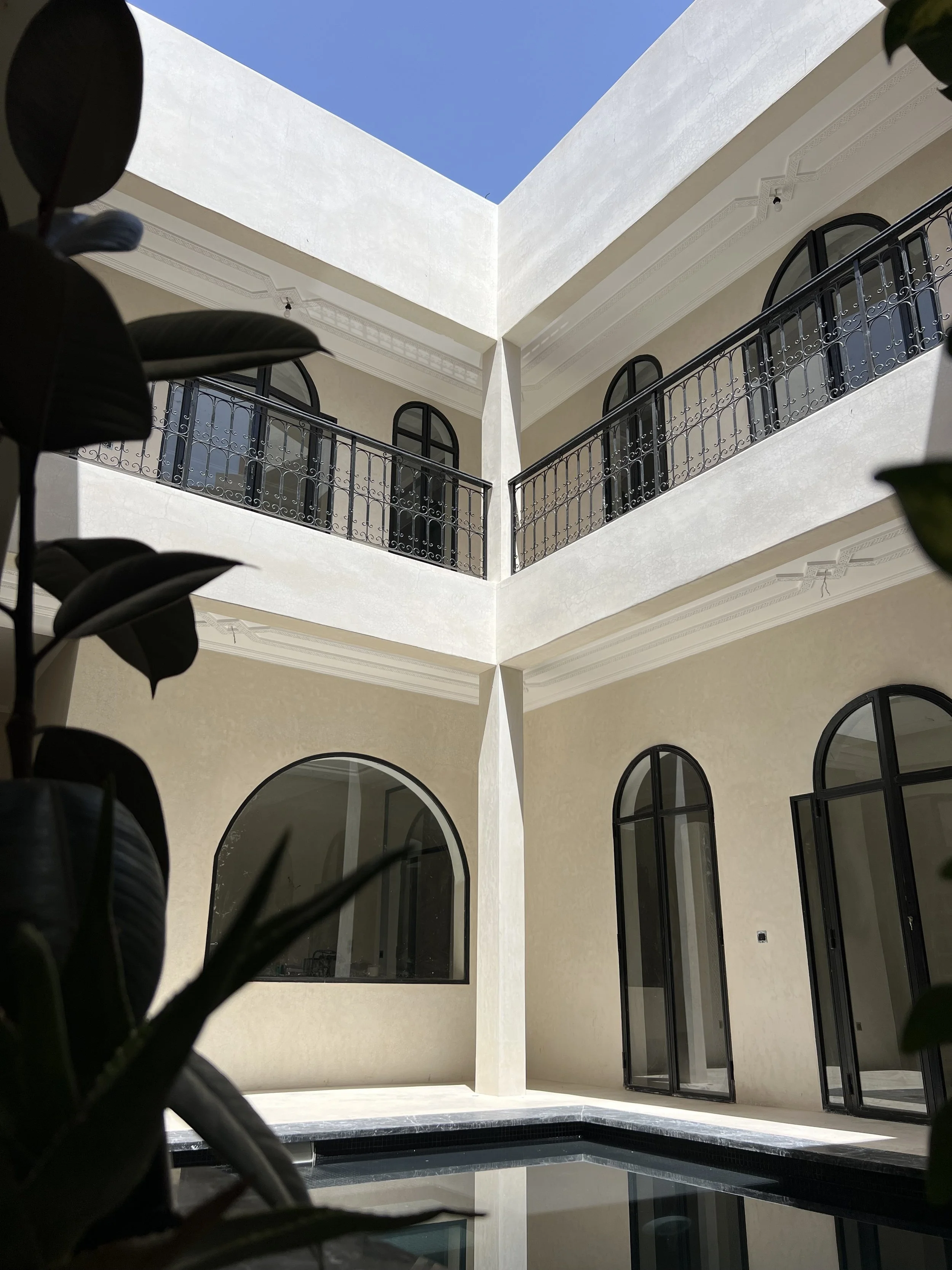 Indoor view of a modern building with tall arched windows, a balcony with black wrought iron railing, cream-colored walls, and a partial view of a swimming pool at the bottom of the image. Blue sky visible at the top.