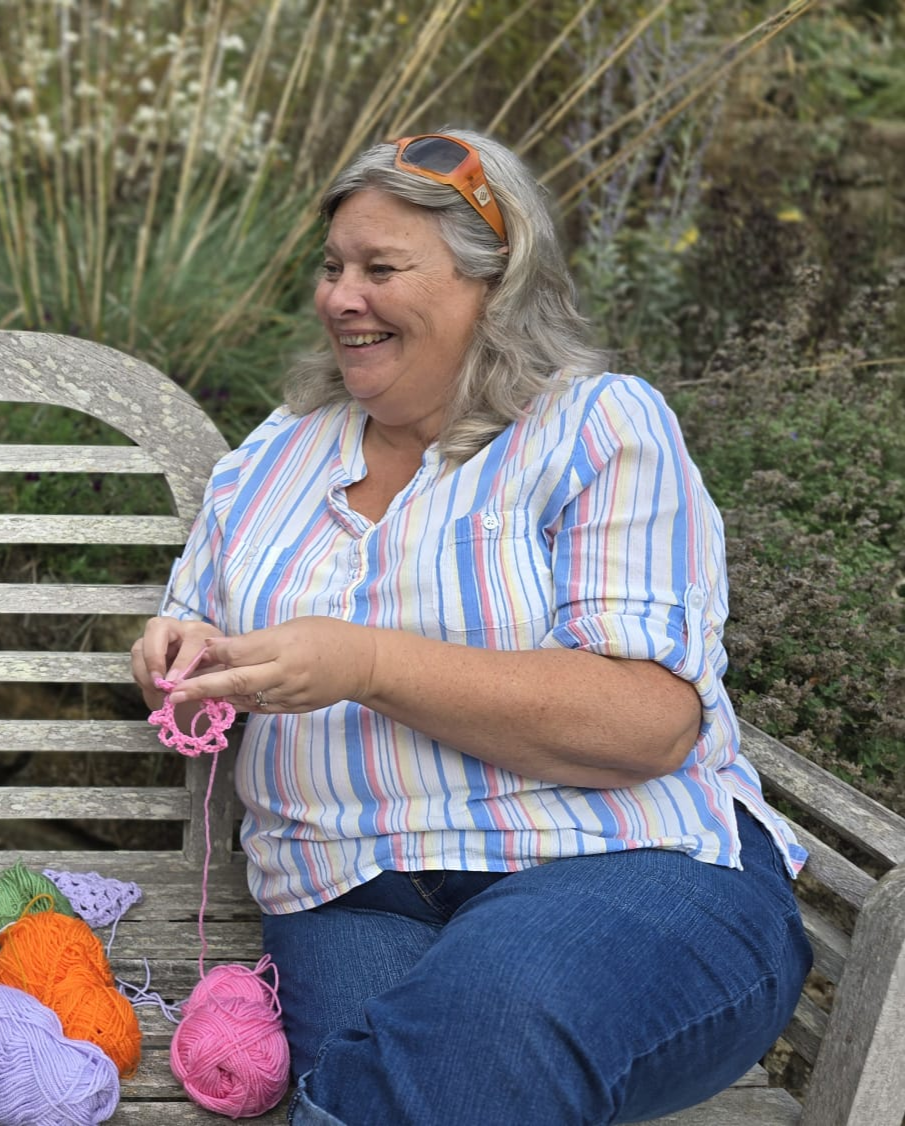 A woman with gray hair wearing sunglasses on her head, a striped shirt, and jeans is sitting on a weathered wooden bench outdoors, smiling and holding pink yarn while surrounded by balls of colorful yarn on her lap and beside her.
