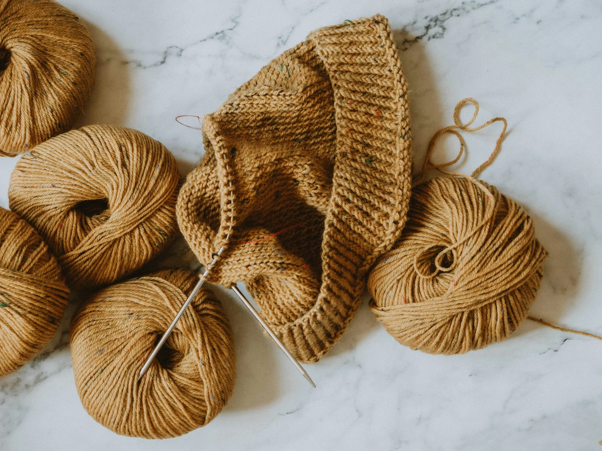 Knitting project on metal knitting needles next to balls of mustard yarn on a marble surface.