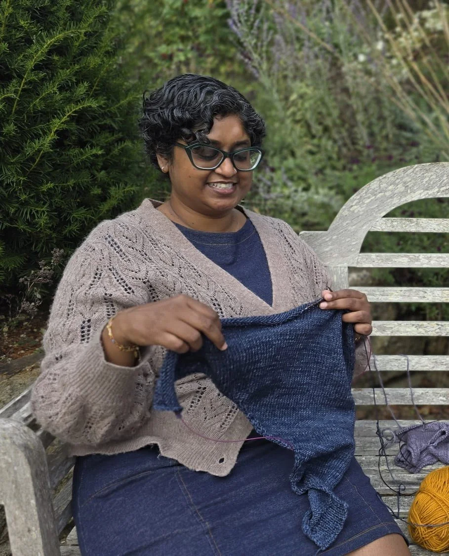 A woman sitting on a wooden bench, knitting with blue yarn outdoors near greenery, smiling while looking at her knitting project.