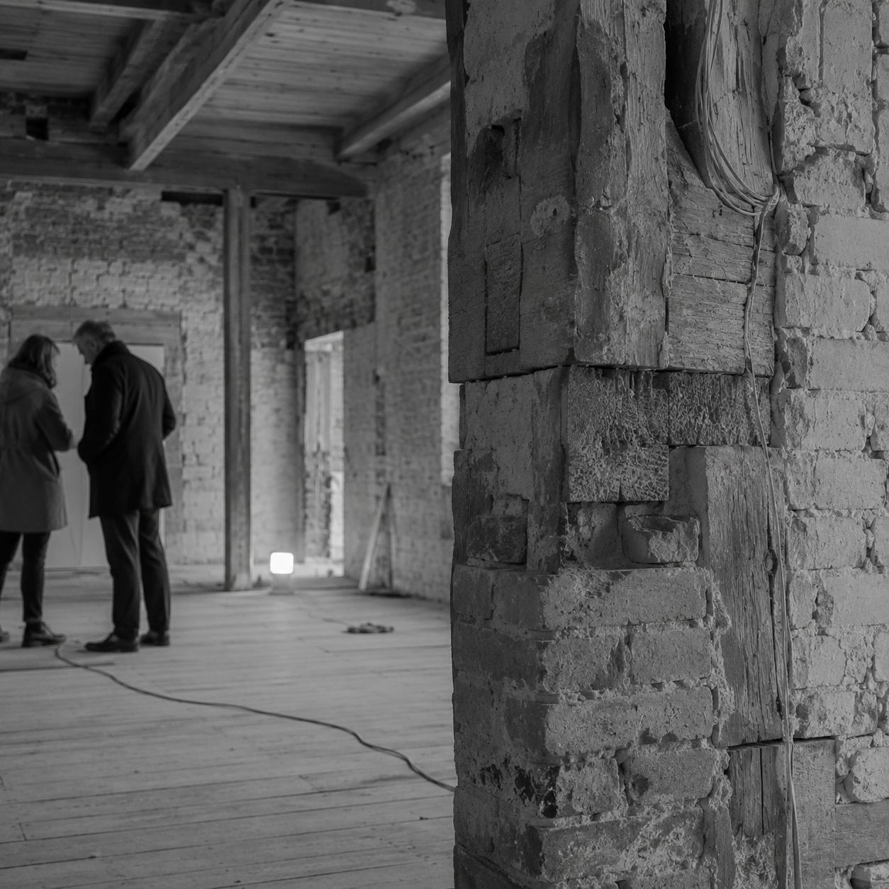 Two people in discussion inside an unfinished brick building with exposed beams, seen from behind a brick pillar.