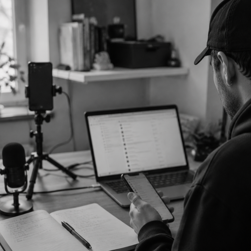 A man with a cap sitting at a desk with a laptop, smartphone, notebook, and pen, recording a podcast with a microphone and webcam in a room.