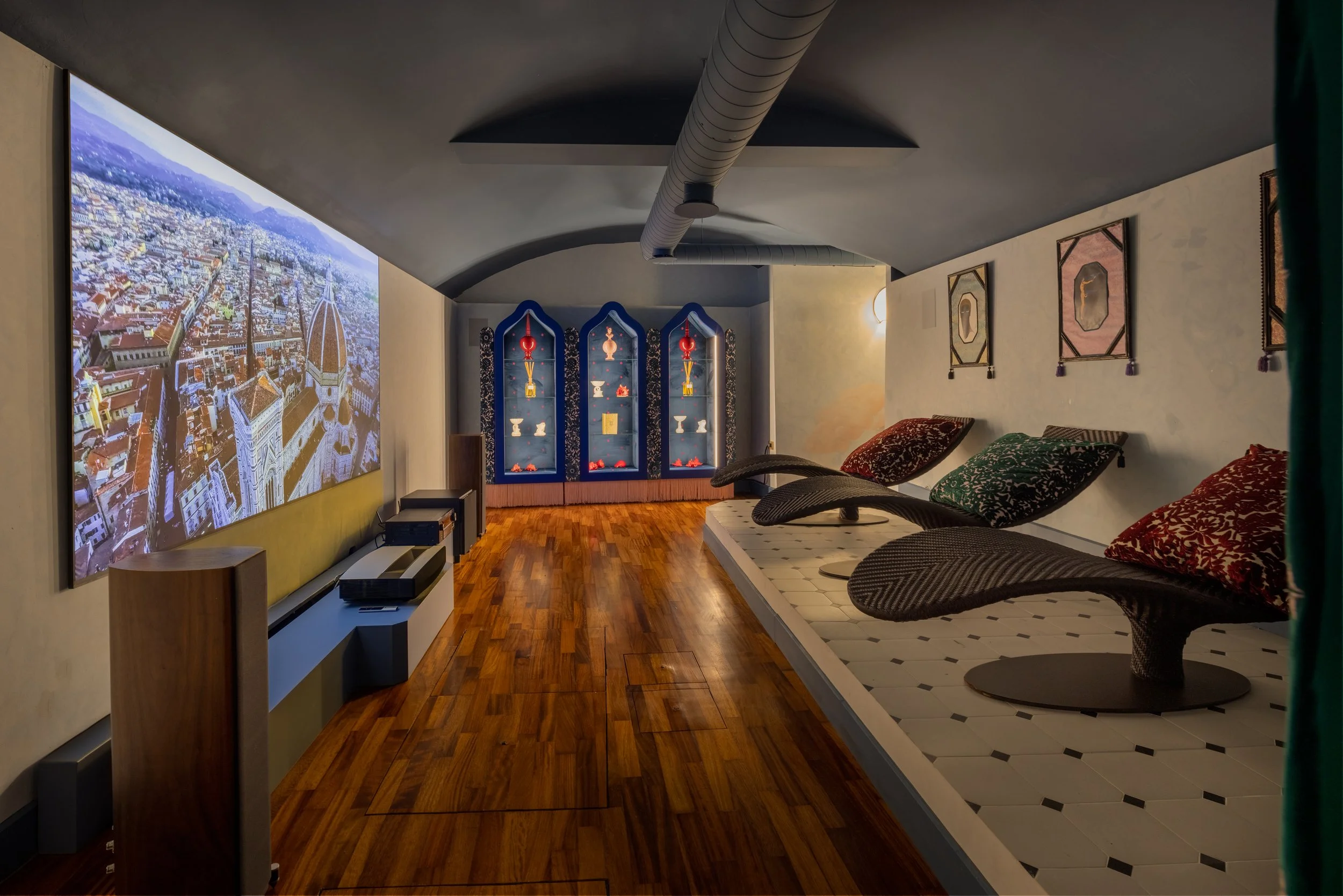 Interno di una stanza con un grande schermo che mostra una vista aerea di una città con montagne sullo sfondo. La stanza ha un soffitto arcuato con un tubo di ventilazione visibile e arredi come una piccola cassettiera, vari cuscini e decorazioni sulle pareti.