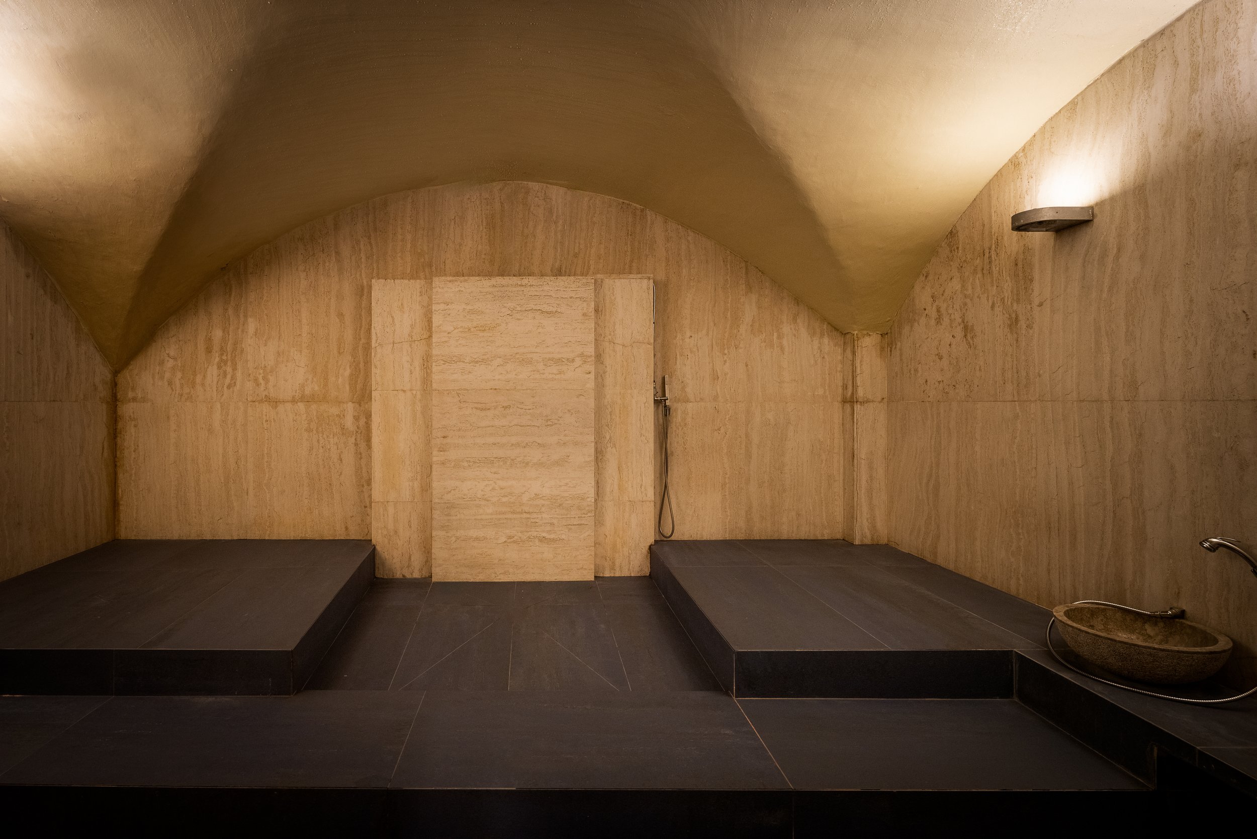 Interno di un bagno turco con pareti di pietra beige, una piattaforma black e un lavandino di pietra con un rubinetto in basso a destra.