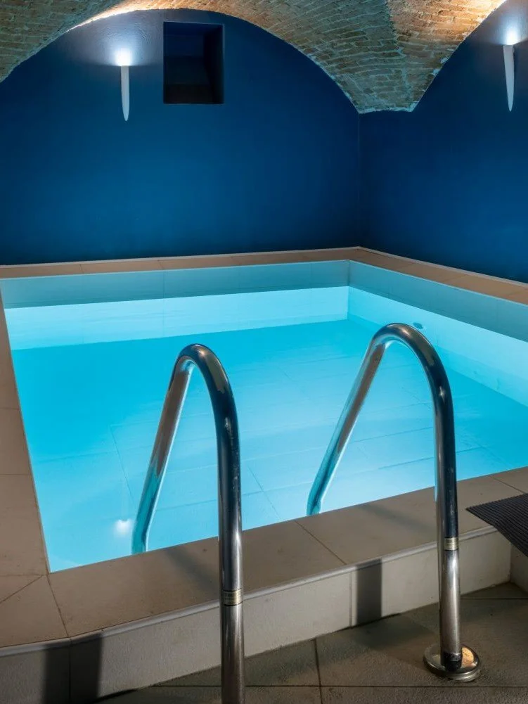 Piscina per il nuoto controcorrente al chiuso con luci soffuse, pareti blu scuro e soffitto in pietra, con scalette in acciaio inox.