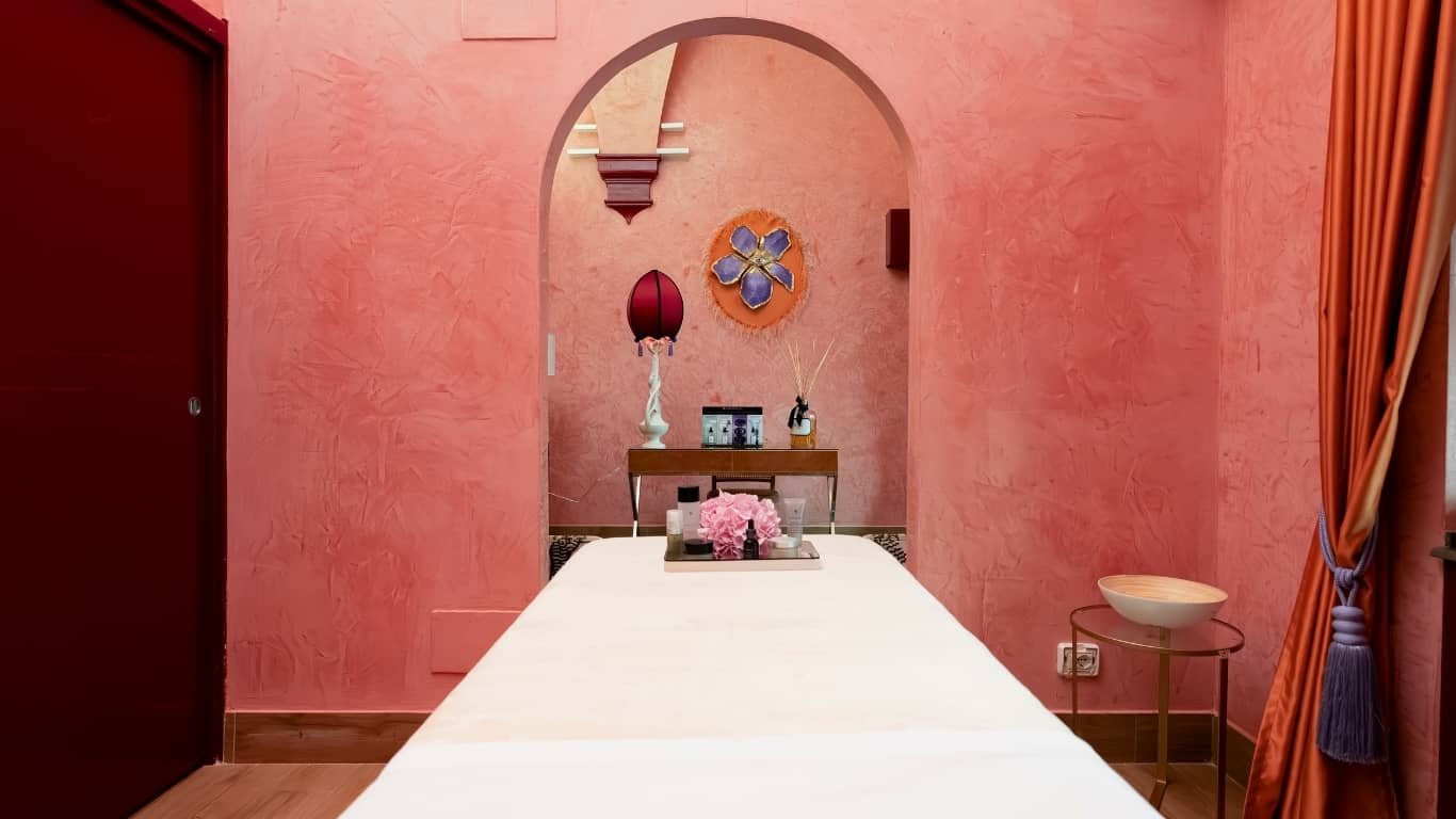 Stanza per massaggi con pareti rosa, tavolo con tovaglia bianca, decorazioni eleganti, lampada rossa, fiore al centro, aria intima e raffinata