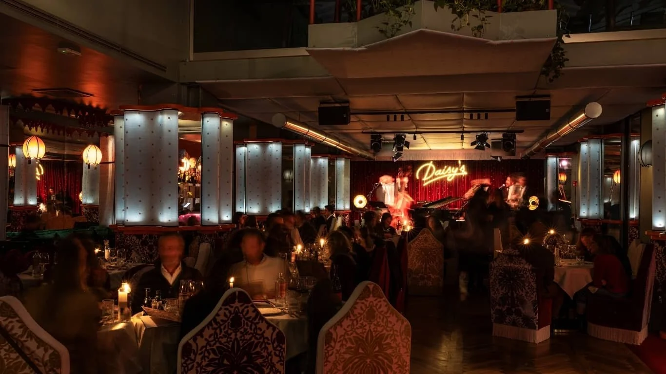 Interno di un ristorante con tavoli apparecchiati, illuminazione soffusa, un palcoscenico con il nome 'Daisy's' illuminato e persone che si gustano la cena, atmosfera vivace e intima.