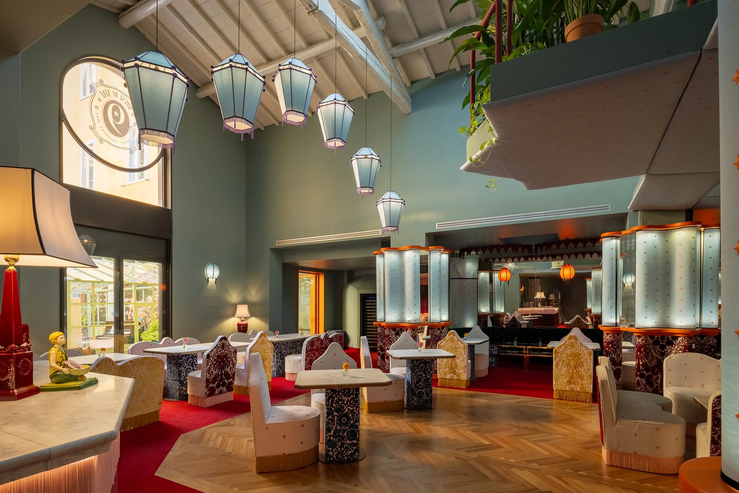 Interno di un ristorante o caffetteria con tavoli e sedie, lampade a lanterna appese al soffitto, pareti verdi, finestra grande luminosa, decorazioni con lampade di diverse forme e una zona con colonne e bar.