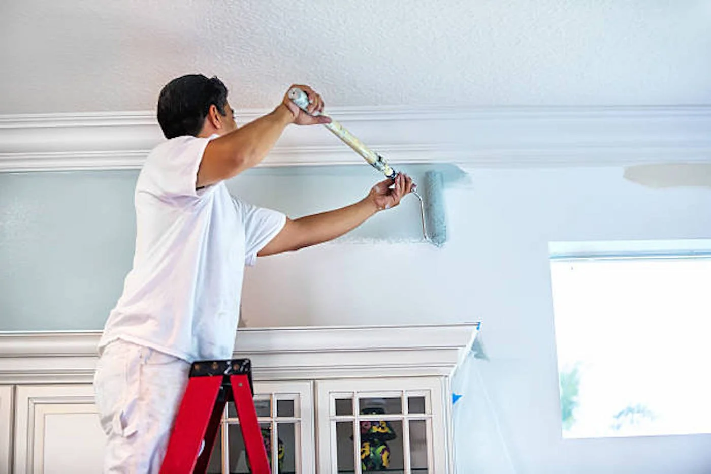 Person painting a ceiling with a paint roller while standing on a ladder.