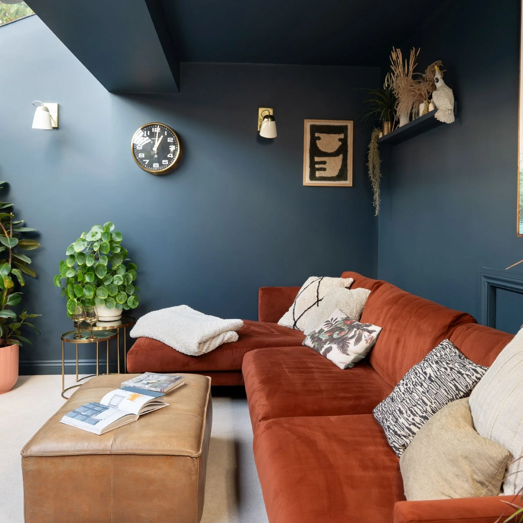 Living room with a rust-colored couch, decorative pillows, a beige ottoman with magazines, a blue wall with a clock, a wall art piece, a plant, and a shelf with decorative items including a bird sculpture.