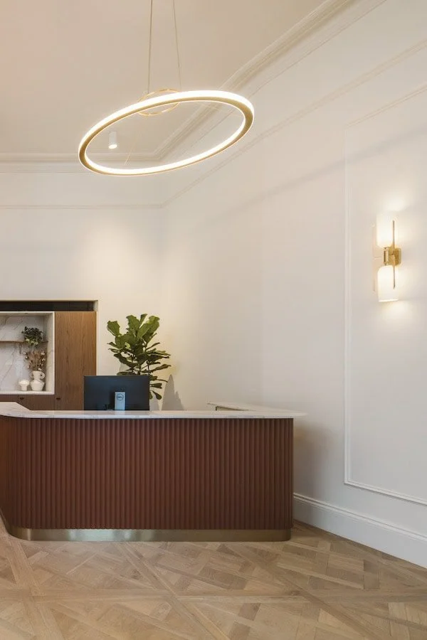 Modern reception desk with a plant and desktop computer, a circular ceiling light, and wall-mounted lighting in a minimalist office or hotel lobby