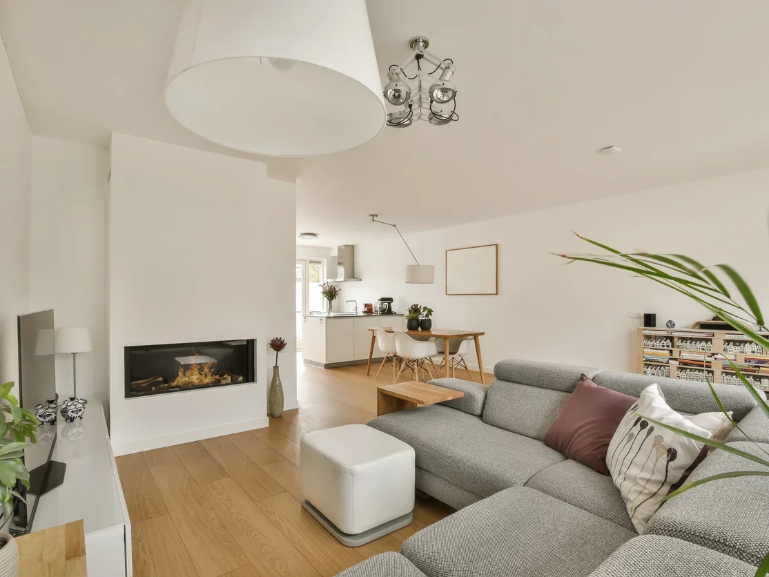 Bright and modern open-concept living room and kitchen with neutral tones, a gray sectional sofa, a wooden coffee table, and a white ottoman. Decor includes pillows, houseplants, and wall art. The space features a built-in fireplace, contemporary lighting fixtures, and a dining area with a round table and chairs. The kitchen has white cabinetry and is illuminated by natural light from the window.