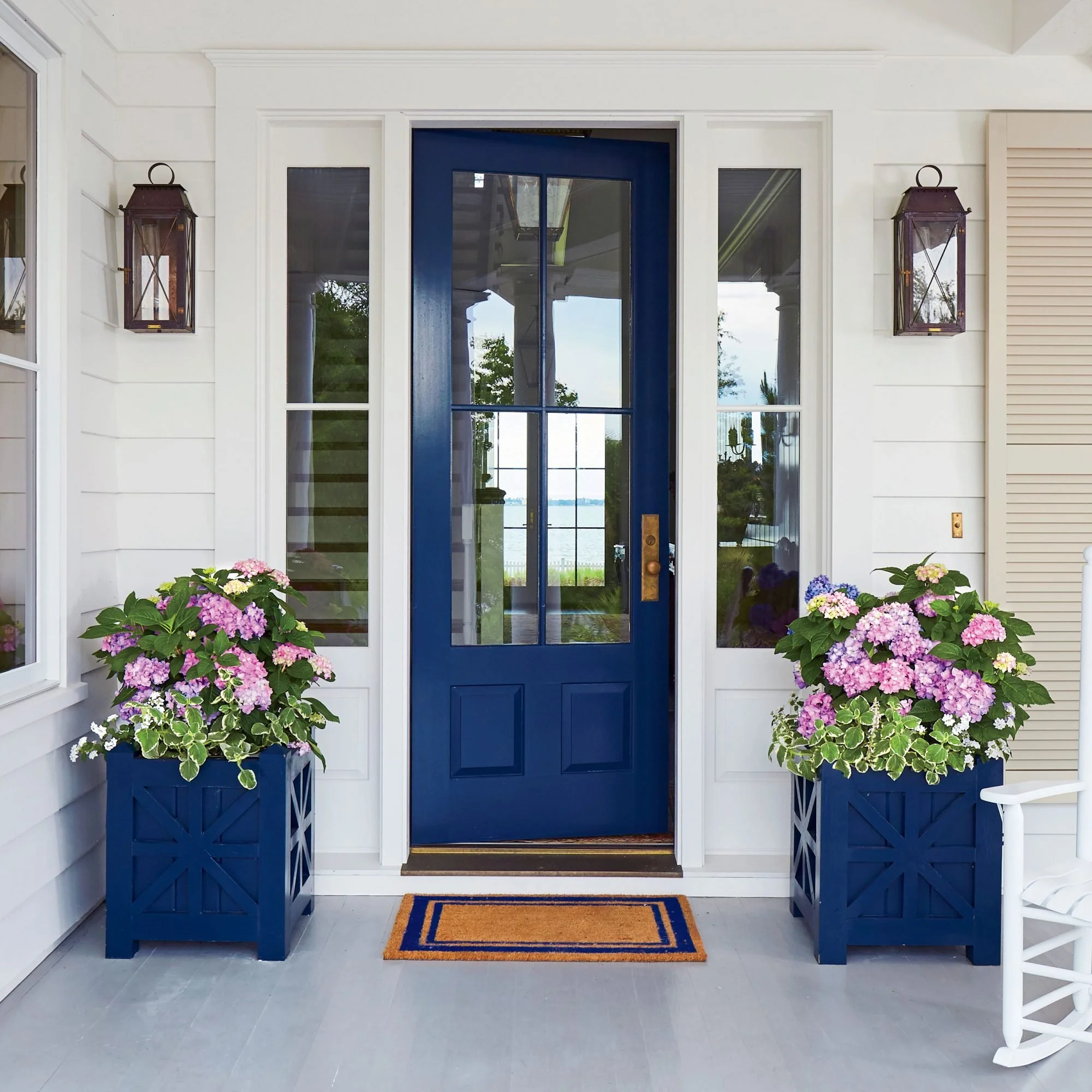 Front porch with a blue door, white framing, two blue planters with pink and purple hydrangeas, and black wall-mounted lanterns on either side.