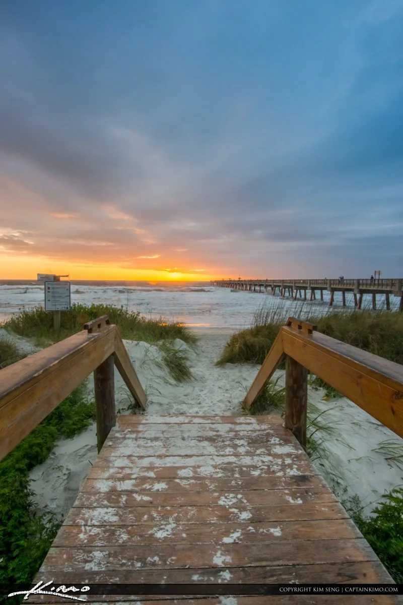 Wooden pathway leading to the beach with ocean waves, a pier extending into the water, and a colorful sunset sky.