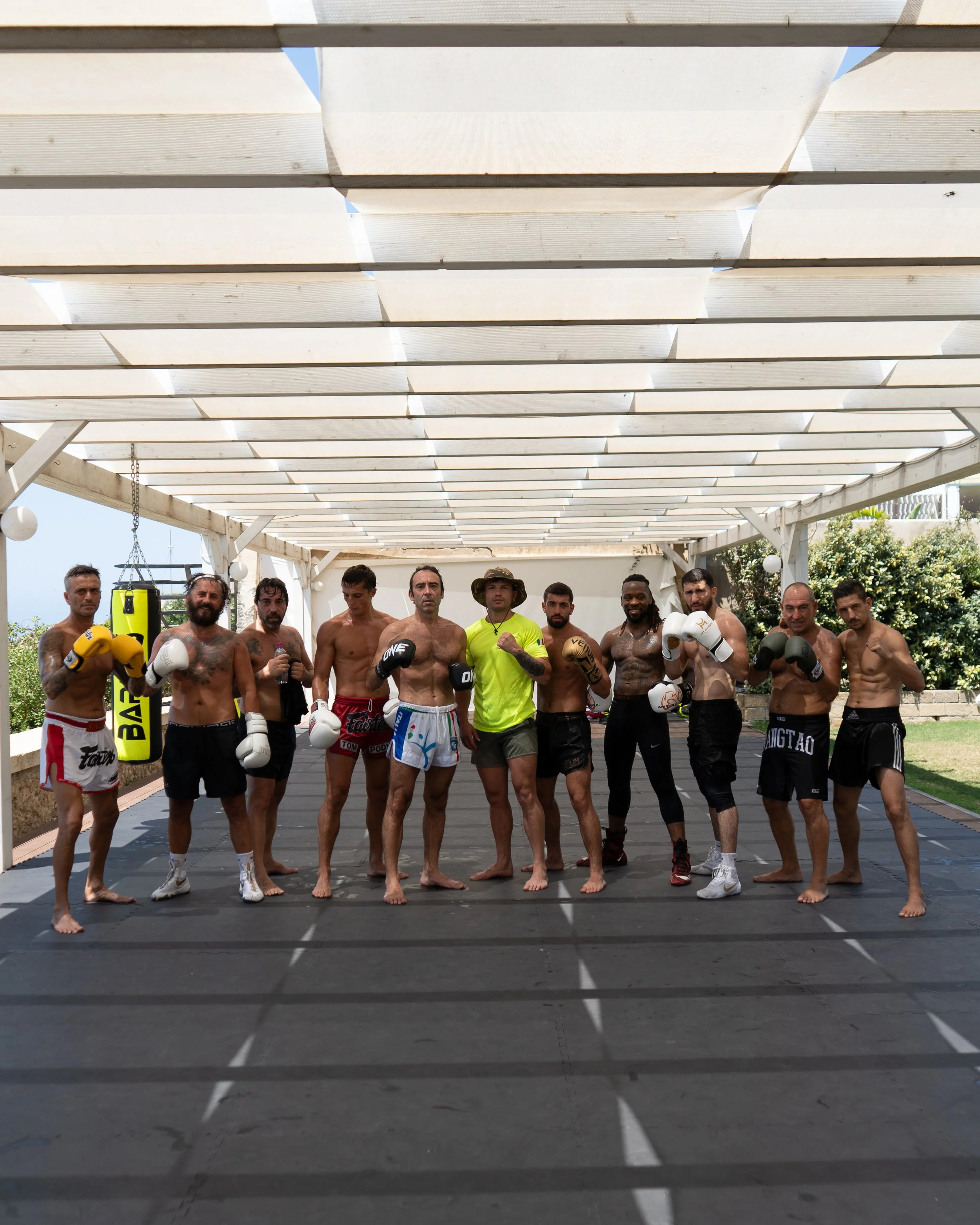 Group of shirtless men in mixed martial arts gloves and shorts, standing in a gym or training area outdoors, under a shade structure with trees and buildings in the background.