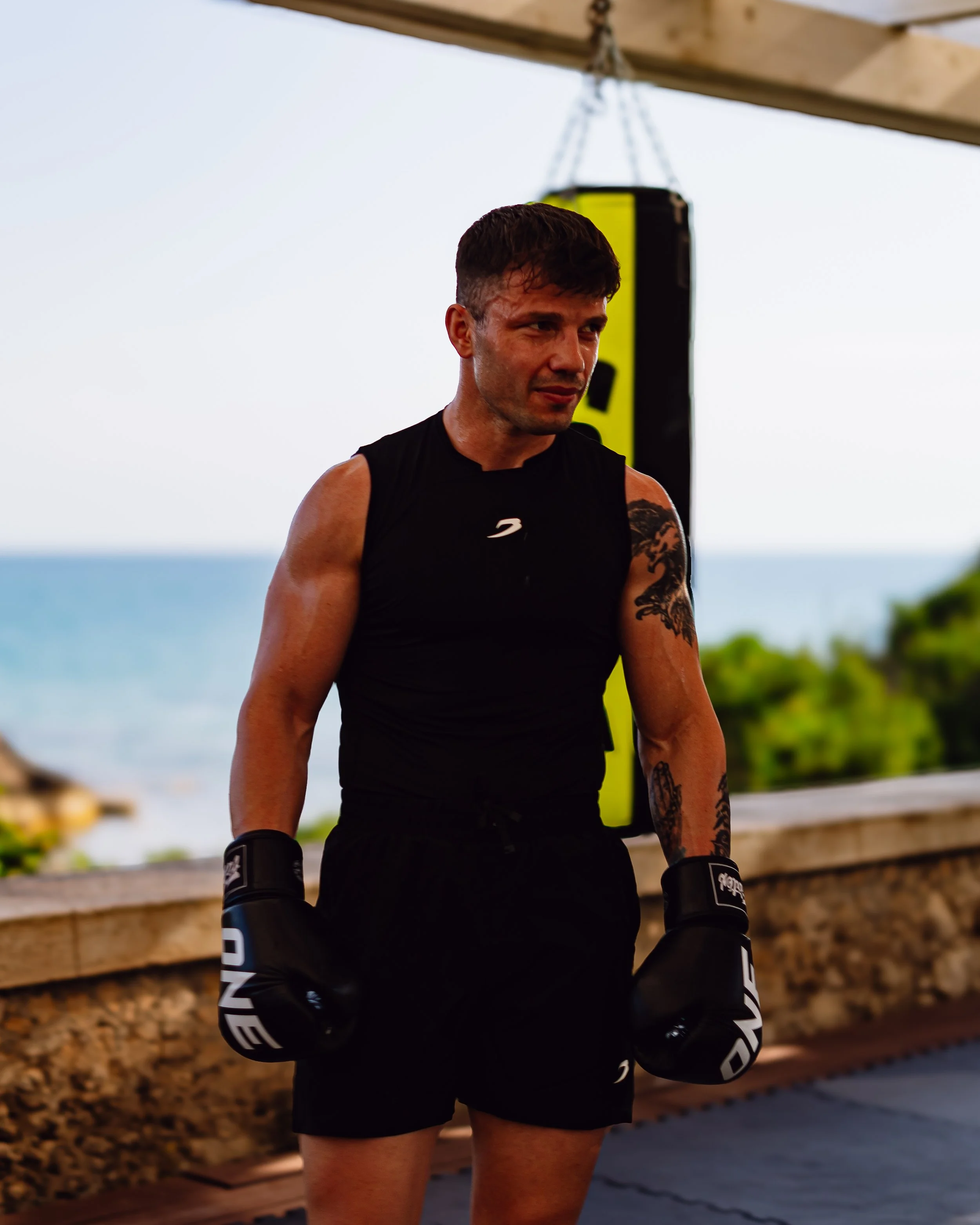 A male boxer wearing black training gloves and black shorts, standing outdoors near a wooden fence with a background of blue sky, trees, and ocean.