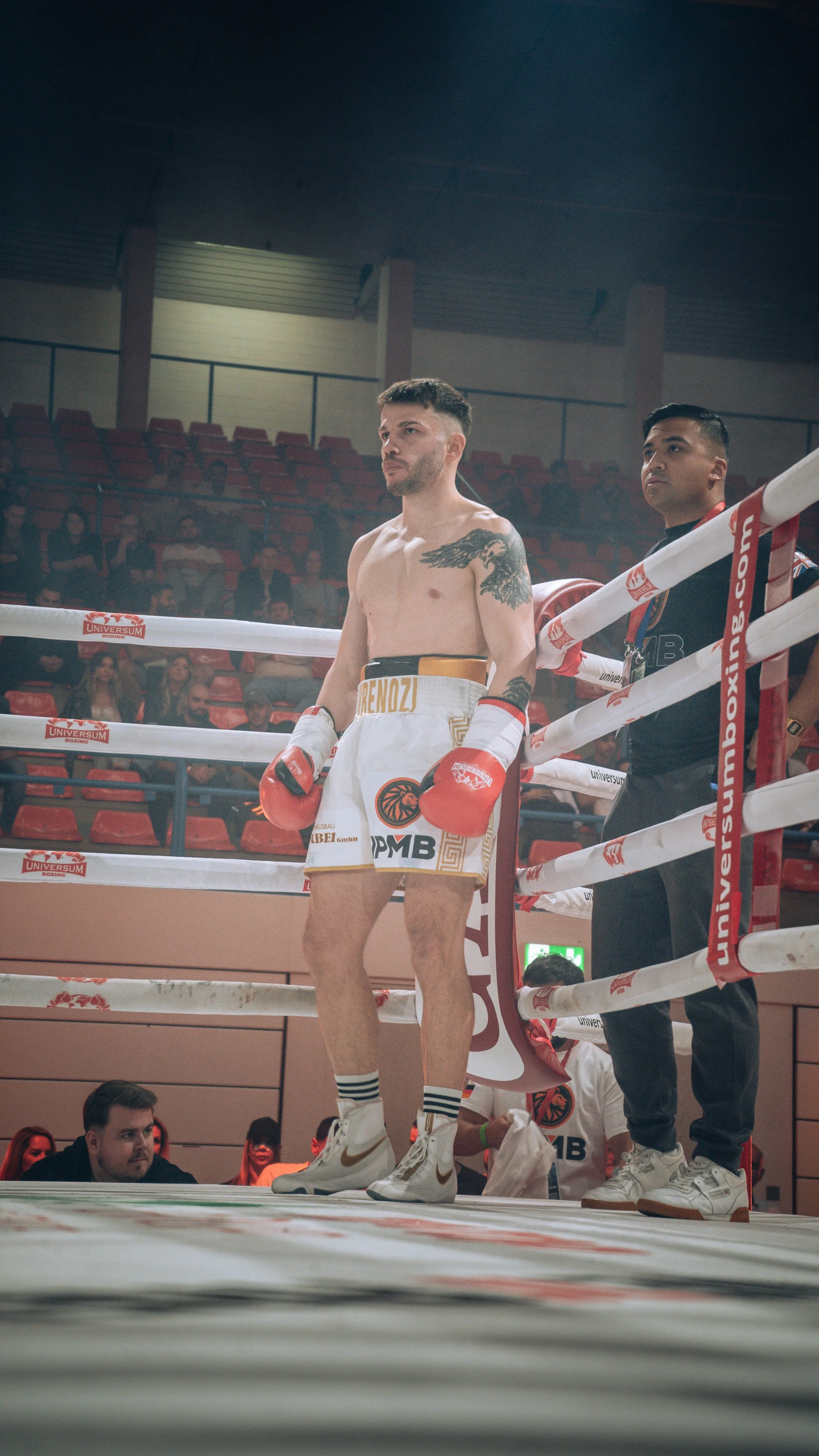 A boxer standing in a boxing ring, wearing white shorts with gold accents and red boxing gloves, with a tattoo on his left shoulder, and a person standing next to him in a dark shirt. Audience members are visible in the background.