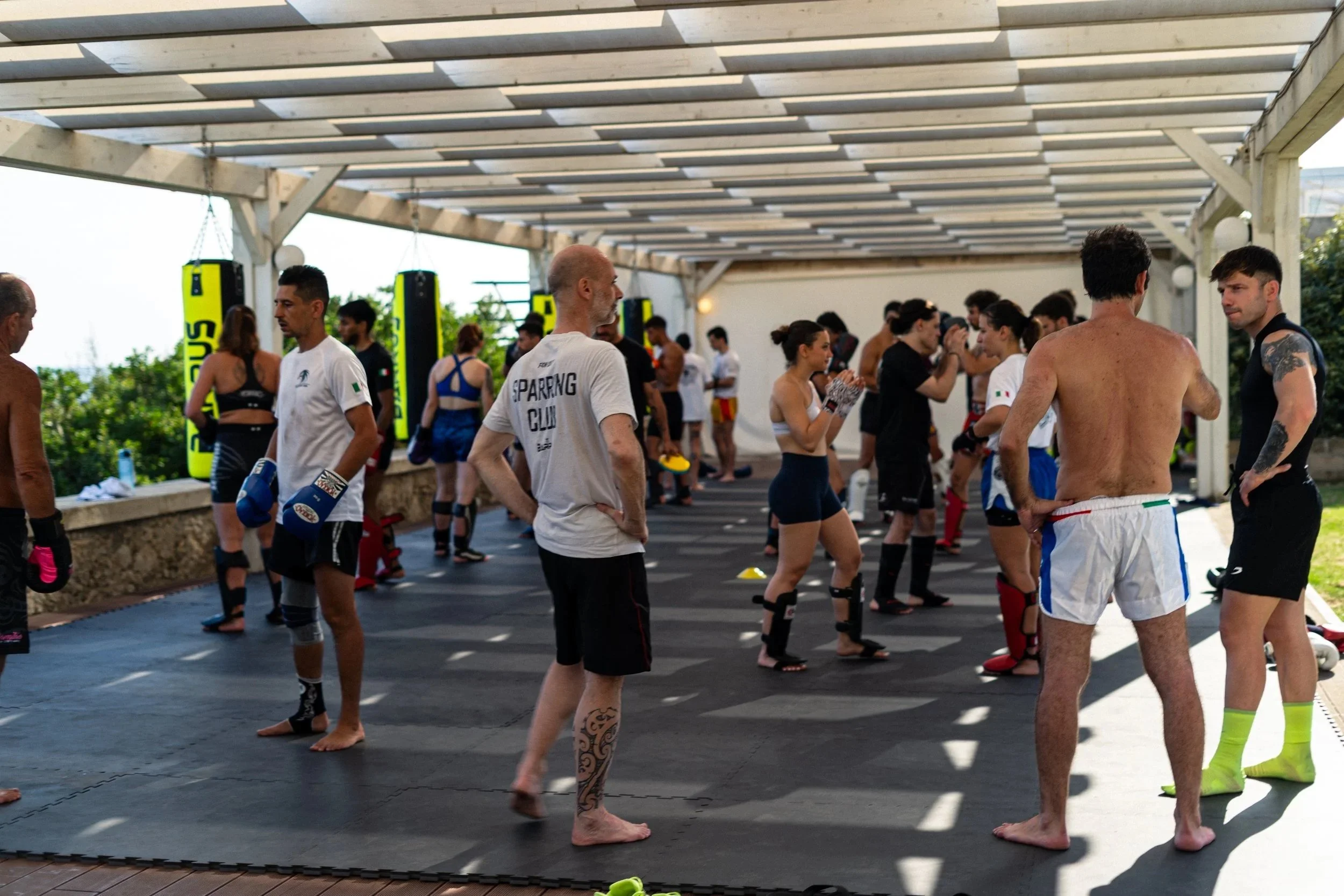 Group of people in athletic wear standing in a shaded outdoor area, preparing for a boxing or martial arts training session, with some wearing boxing gloves.